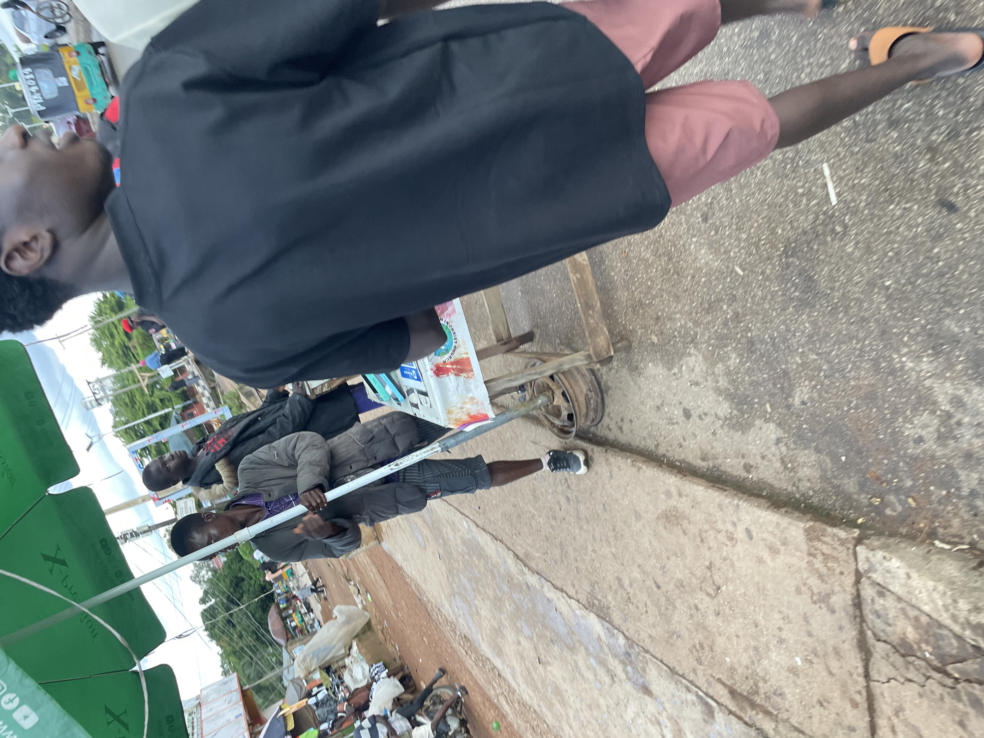 In the city of Jos, Nigeria, on a street likely bustling with activity, a group of individuals are captured in a candid moment. The foreground is dominated by the back of a person wearing a black t-shirt and pink shorts, their stride suggesting movement. To the left, a man in a gray jacket and shorts stands under a large green umbrella, holding a white pole. Beside him, another man in a dark jacket is partially visible. The background reveals a typical street scene in Jos, with market stalls, a motorized tricycle (keke napep) with license plate PTA 4011, and other pedestrians. The ground is paved with concrete and asphalt, with some areas appearing wet. The overall impression is of a lively street environment, perhaps a marketplace or a transit point, where daily life unfolds.