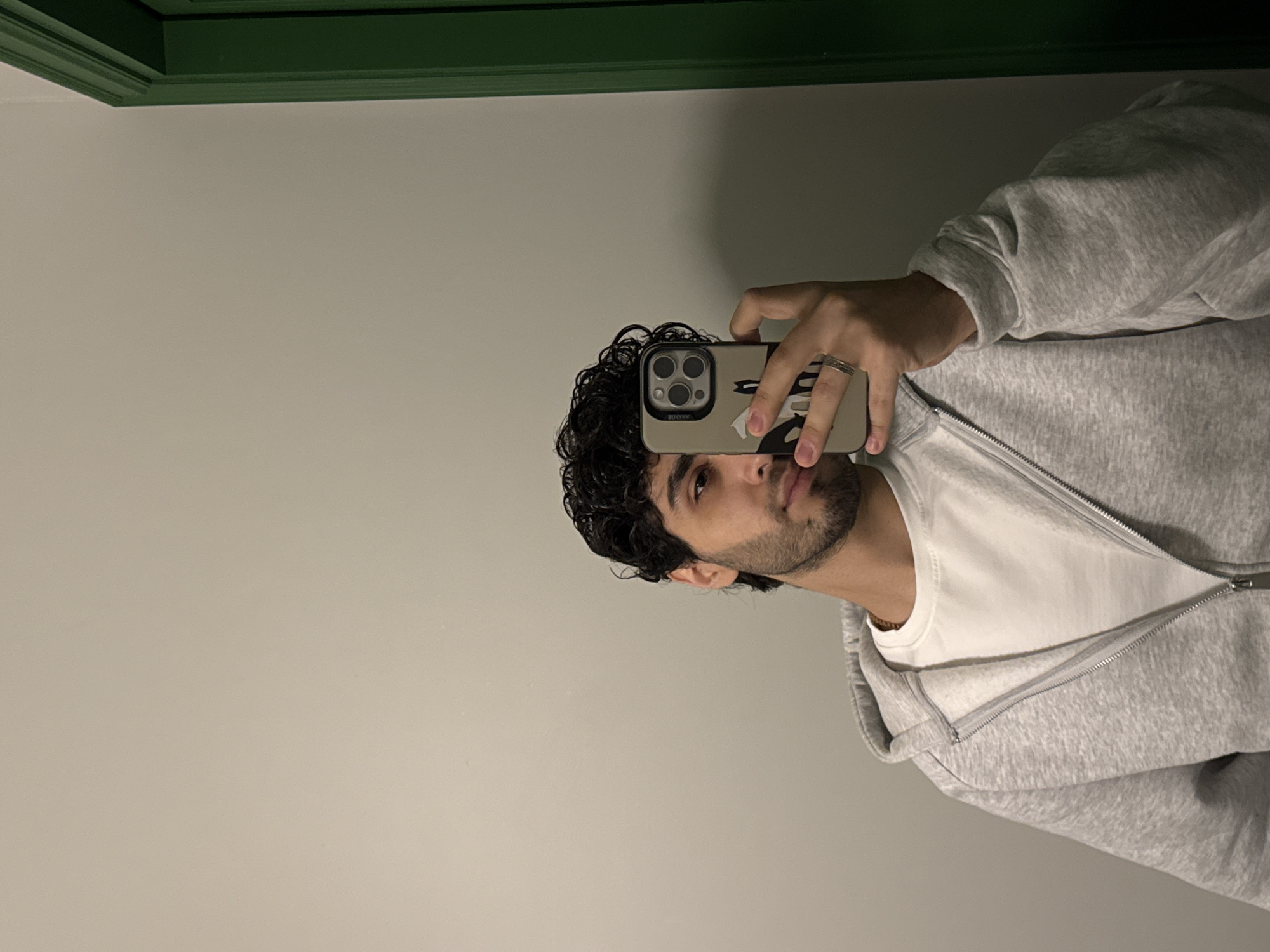 A man is holding a cell phone to take a picture. He is wearing a gray hoodie over a white t-shirt. The background is a light gray wall with green trim at the top. There is no evidence to suggest that the image was taken in Yerevan, Armenia.
