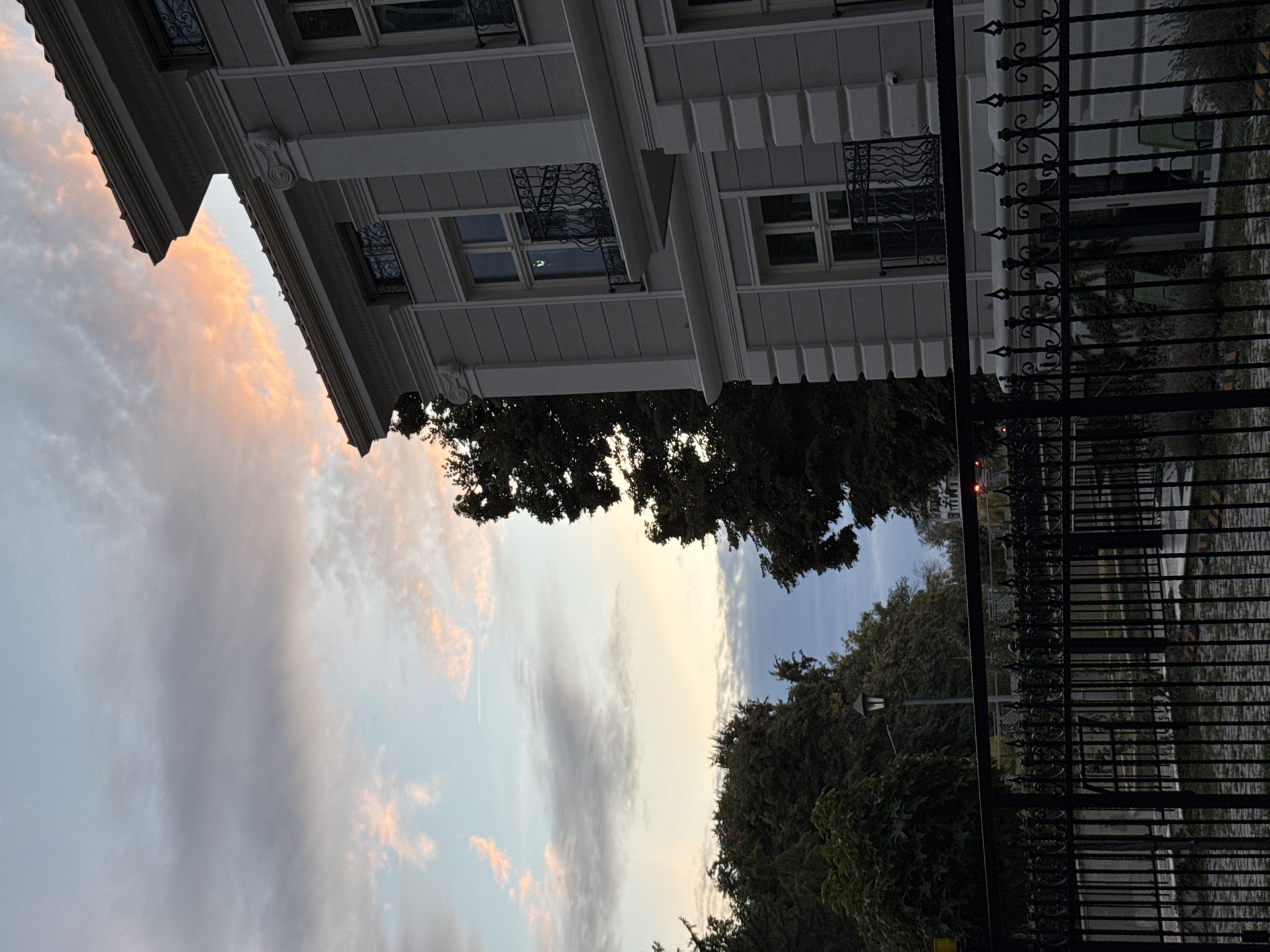 The image depicts a facade of a building with neoclassical architectural elements, including ornate columns and window embellishments, set against a twilight sky with wispy clouds tinged with orange and pink hues. A dark metal fence runs across the foreground, partially obscuring a cobblestone pathway and glimpses of greenery and a street lamp. The building appears to be a residential or commercial property. Based on the architectural style and the quality of light, this scene is consistent with the area around Tsar Simeon's Garden or the central pedestrian streets of Plovdiv, Bulgaria, particularly during the evening or early morning hours. There are no visible people in the frame, and the environment suggests a calm, urban setting.