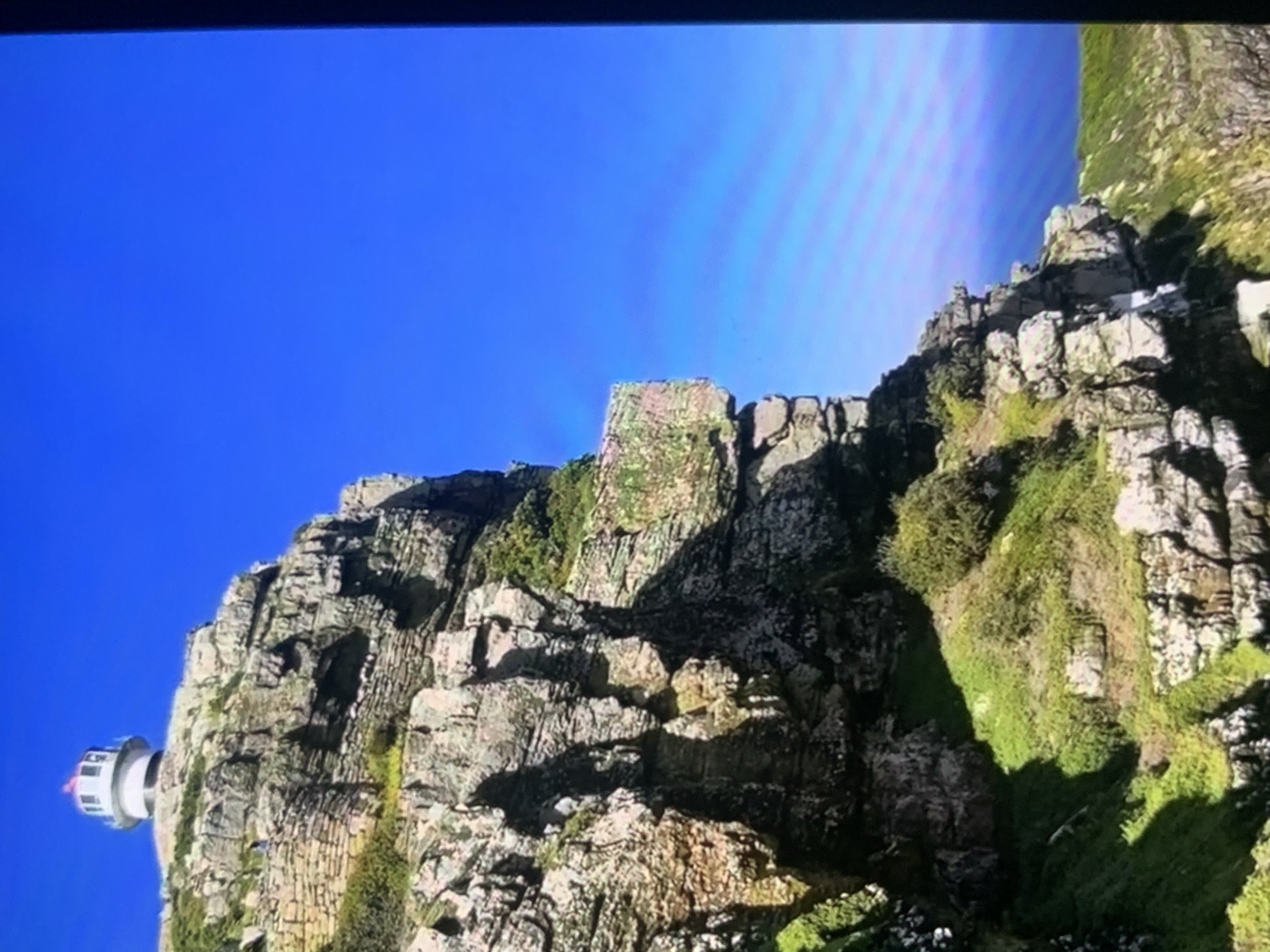 A lighthouse stands atop a rocky cliff overlooking a body of water, bathed in bright sunlight under a clear blue sky. The cliff face is rugged and uneven, with patches of green vegetation clinging to its surfaces. The lighthouse is white with a red-capped lantern room. The scene is devoid of any people. The image does not depict any location in the city of Stavropol, Russia.