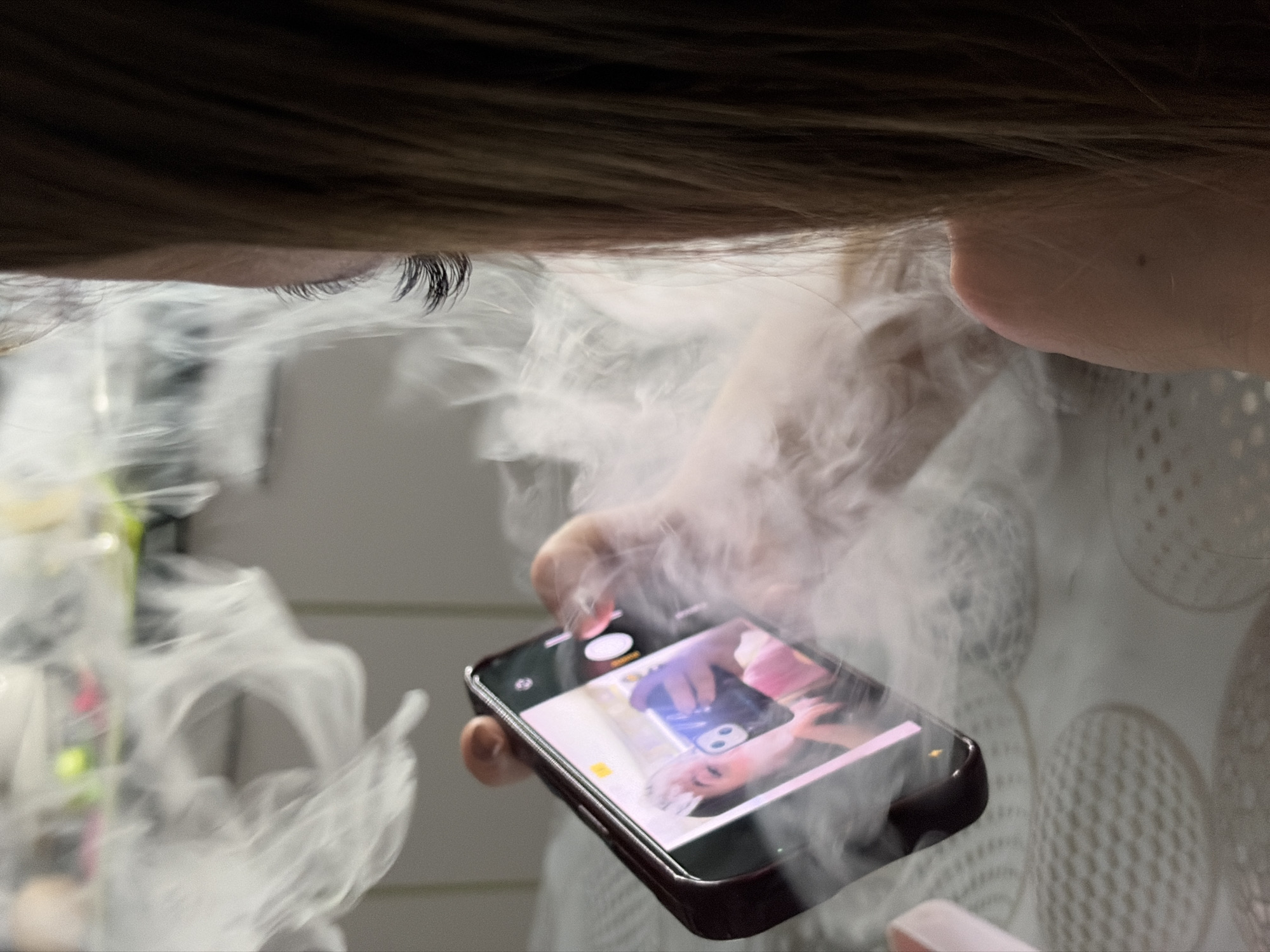 A close-up shot depicts a person's face and hand holding a smartphone engulfed in white smoke. Only the upper part of the face is visible, including the eyelashes and forehead beneath dark hair. The person's hand is holding a black smartphone, with the screen displaying a photograph. The scene is obscured by a thick cloud of white smoke, which partially obscures the background. The background appears to show some indistinct shapes and patterns, possibly fabric or furniture. The location, Port-Katon, Russia, is not discernible from the visual information provided.