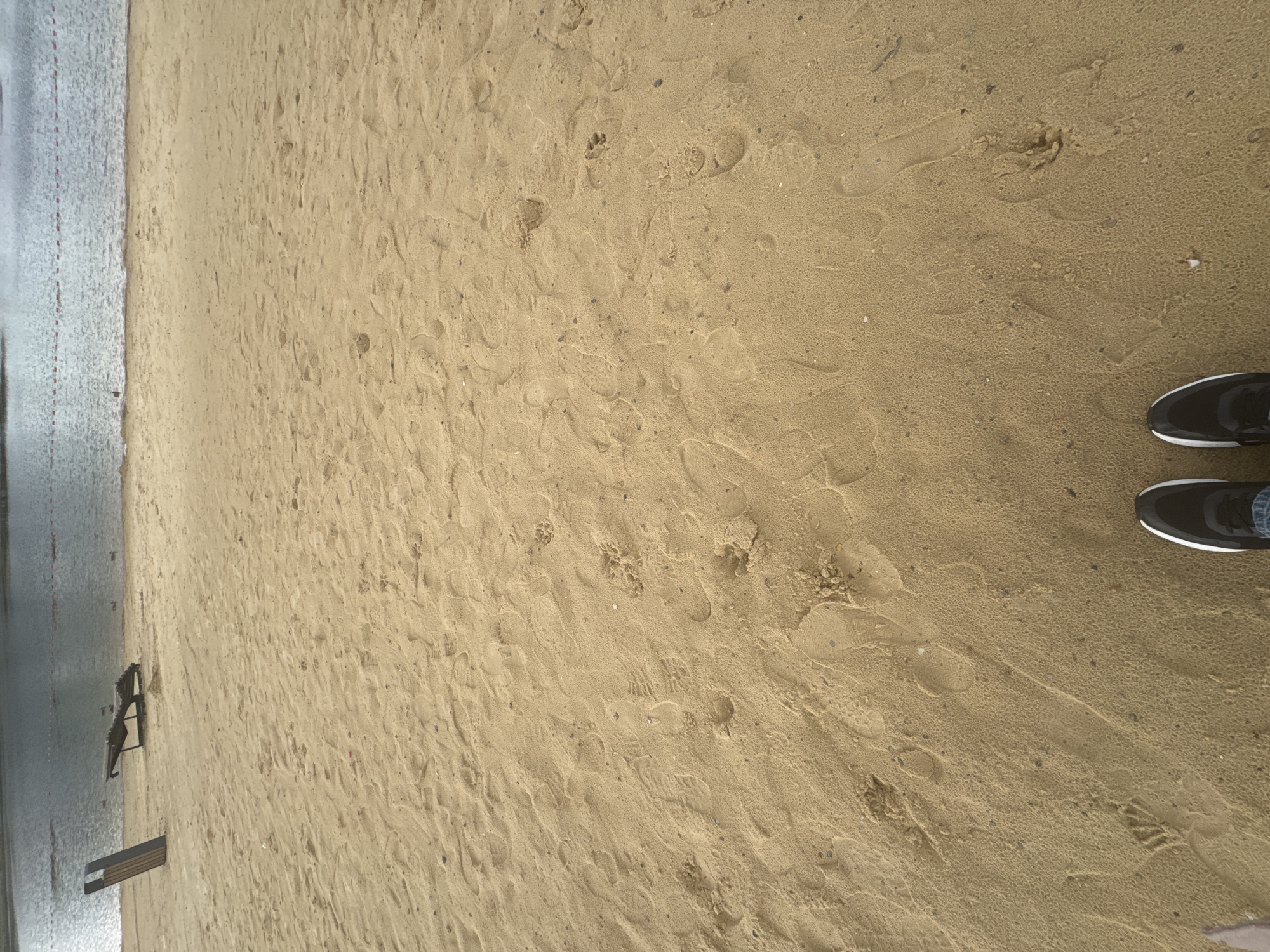 The image depicts a sandy beach with footprints visible on the sand. In the background, a body of water is present, with some buoys marking a boundary. Two lounge chairs and a trash can are also visible on the sand. The foreground shows a person's feet wearing black and white sneakers. The overall impression is of a serene, possibly overcast day at a recreational area. The location is the beach of the Ufa Lake in Ufa, Russia.