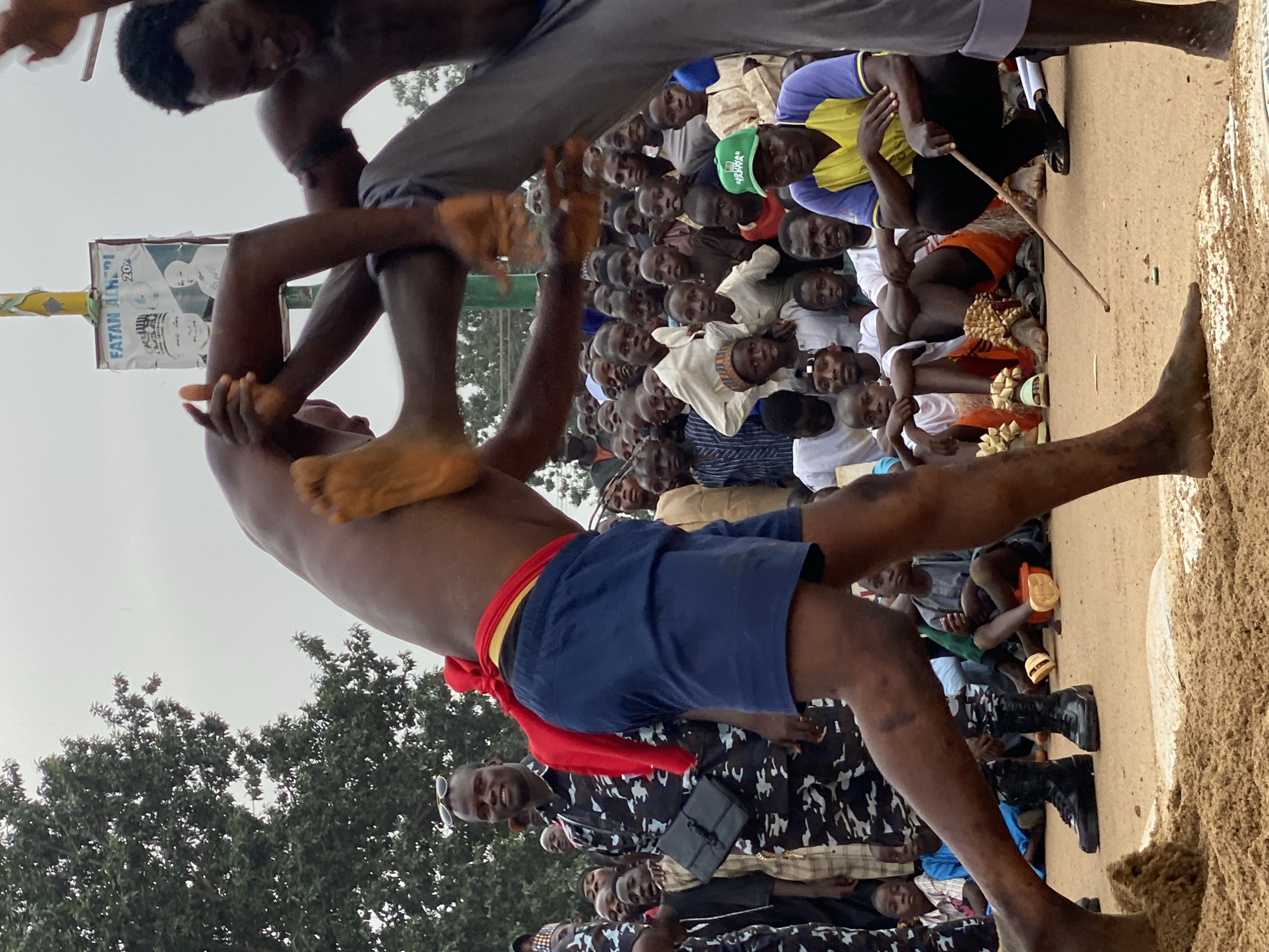 Two shirtless men are wrestling in a sand pit surrounded by a large crowd of onlookers in Kafanchan A, Nigeria. One wrestler wears blue shorts with a red sash, while the other wears grey pants. A poster hangs from a pole behind them.