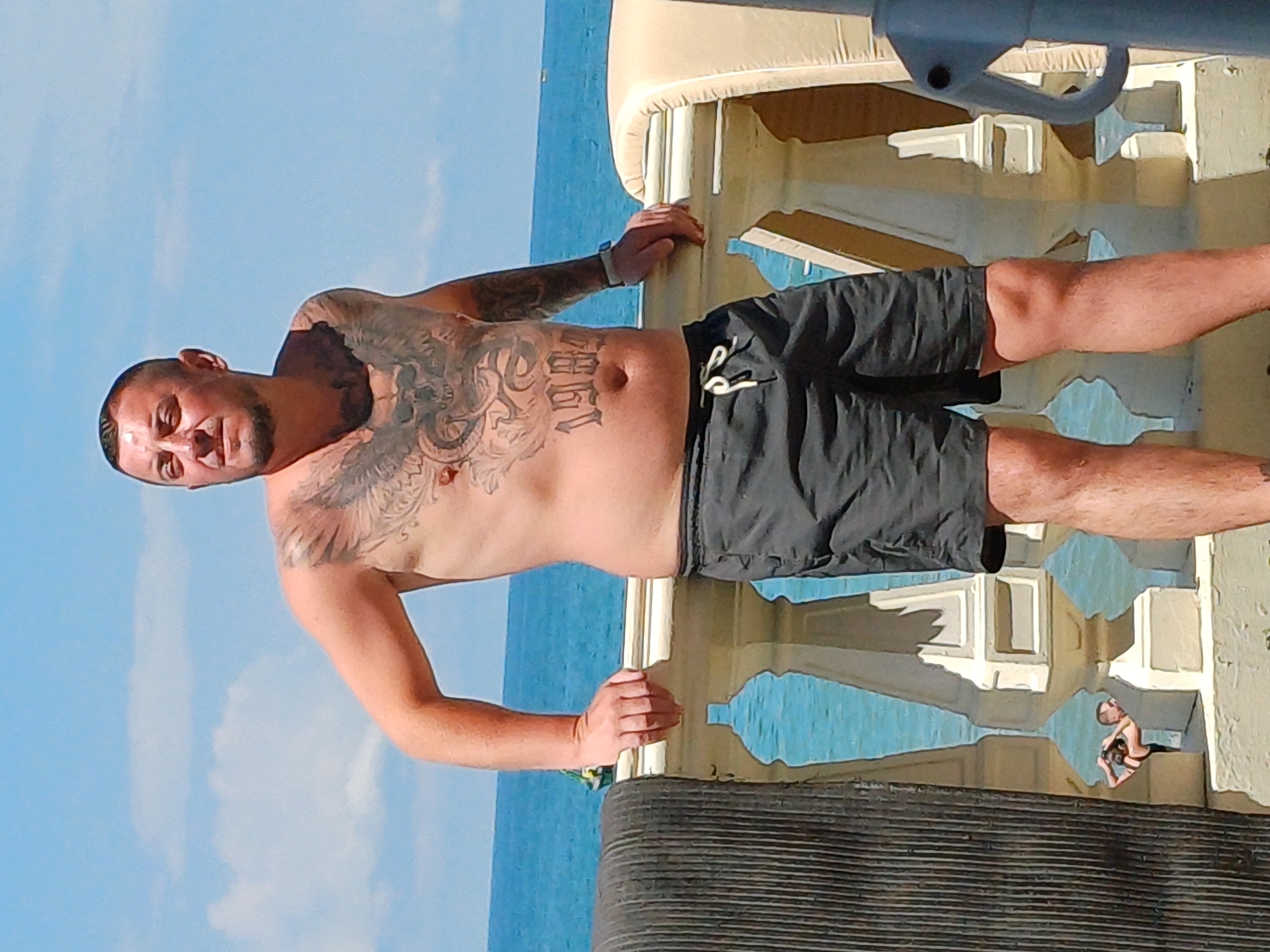 A man with tattoos on his chest and arms, wearing black shorts, stands on a balcony overlooking the Black Sea. He is looking to his left. The balcony has a white ornate railing. In the background, the turquoise sea meets a blue sky with scattered clouds. Two small figures are visible in the water below the balcony. The overall scene is a sunny day, likely in Sochi, Russia, given the context.