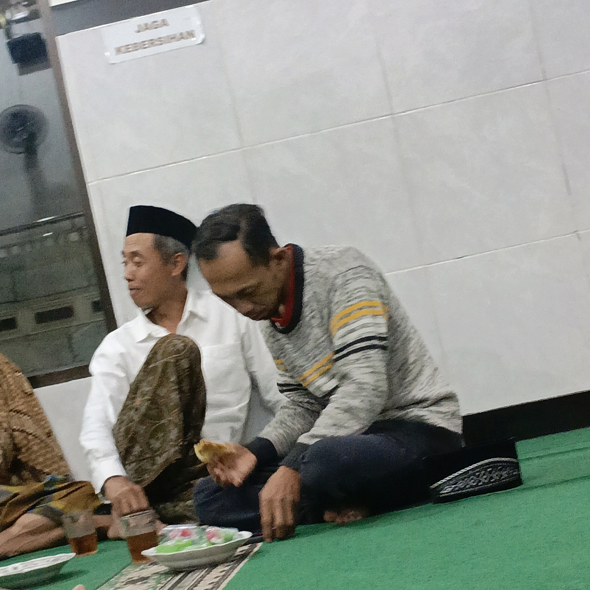 The image shows two men sitting on a green carpet. The man on the left is wearing a white shirt and a black peci (a cap commonly worn by Muslim men in Indonesia). He is looking to his left and appears to be in profile. The man on the right is wearing a grey sweater with yellow and black stripes and dark pants. He is looking down and appears to be holding a piece of food in his right hand. In front of them, there are plates with food and cups of drinks. A sign with Indonesian text "JAGA KEBERSIHAN" which means "MAINTAIN CLEANLINESS" is visible on the wall in the background. The environment suggests an indoor setting, possibly a mosque or a community gathering place in Salatiga, Indonesia.