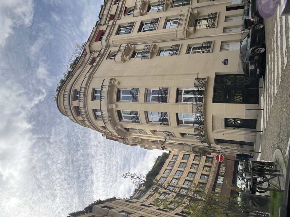 The image depicts a street corner in the 16th arrondissement of Paris, France, at the intersection of Rue Paul Nicaud and Rue du Général Niox. The scene is observed under an overcast sky featuring scattered, textured cloud formations.
A prominent five-story corner building, constructed of light beige stone in a Haussmannian architectural style, occupies the right and central portions of the frame. This building exhibits a curved facade at the corner, multiple windows, and various black wrought-iron balconettes and balconies. The upper stories feature a continuous, recessed balcony with retracted red awnings and visible rooftop greenery. The ground floor includes two large dark metal double doors, an ornate single entrance door on the curved corner, and a blue street sign identifying "Rue Paul Nicaud."
To the left, a multi-story building with a curved, beige facade and rectangular windows is partially visible, also featuring extensive greenery on its rooftop.
The street is paved with cobblestones, flanked by a paved sidewalk with black metal bollards. A marked pedestrian crosswalk is visible in the foreground. A red circular "No Entry" traffic sign is positioned on the left side, near a small, sparsely-leafed tree. A black metal bike rack is partially visible in the lower left.
A black Polestar 2 sedan, with a partially visible French license plate "FH-486-EJ," is parked on the street to the right. A silver/grey car is parked directly behind it, partially obscured. Additional parked vehicles are visible further down the street on the left. Two indistinct human figures are walking away from the camera on the left sidewalk, near the "No Entry" sign. A street lamp is affixed to the main corner building.