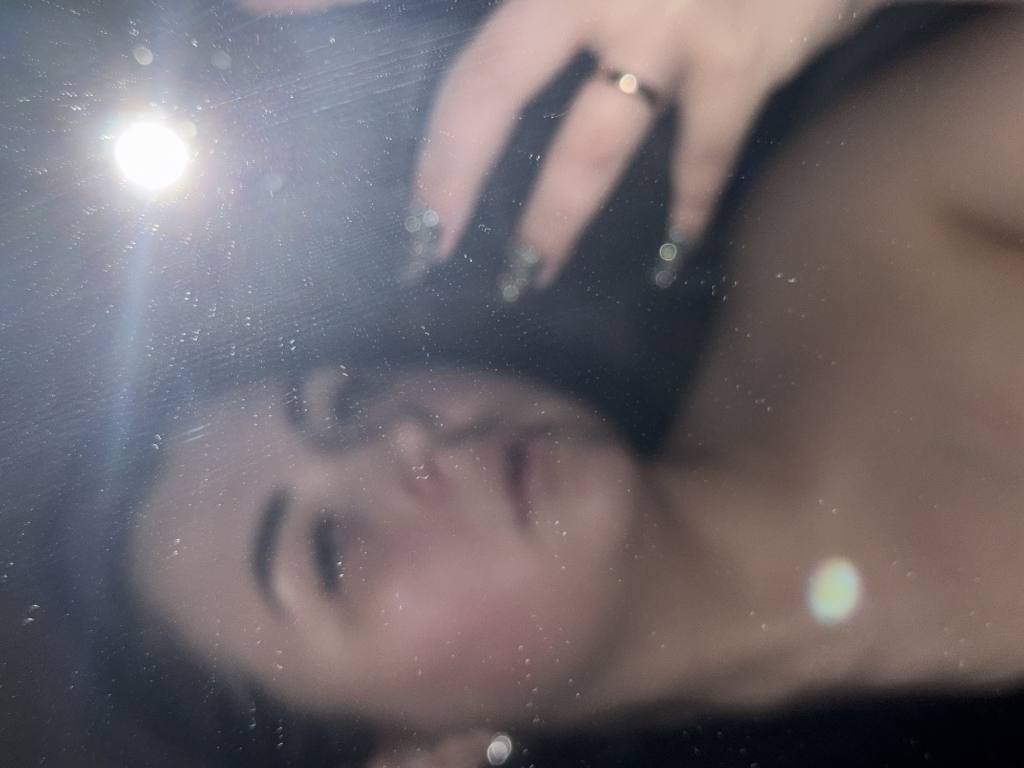 A blurred image of a woman's face is visible through a textured or dirty transparent surface, possibly glass. A hand with a ring on one finger and dark nail polish is visible above the face. A bright light source is present, causing lens flare and further obscuring the image. No specific location in Chaikovsky, Russia can be determined from this image.