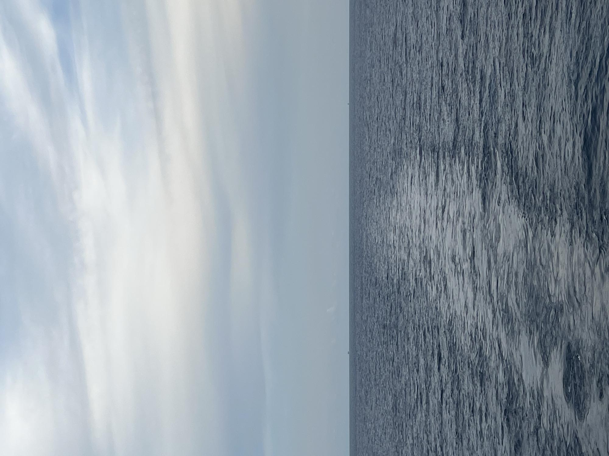 A vast expanse of dark, choppy ocean stretches across the lower half of the frame, meeting a pale, overcast sky at the horizon. Wispy clouds, tinged with a light blue, drift across the sky. The water's surface is rippled and textured, with a subtle sheen of light reflecting off its waves. In the distance, a tiny, dark rectangular shape, possibly a distant vessel or structure, is barely visible on the horizon line, suggesting a seascape rather than an urban environment. The overall impression is one of open space and a muted, tranquil atmosphere.