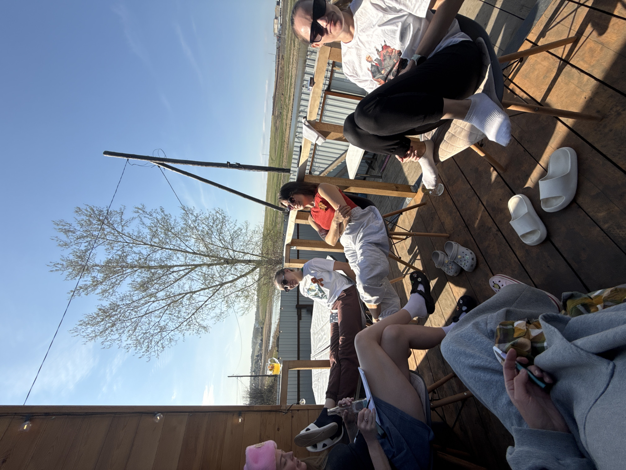 A group of people are gathered on a wooden deck outdoors. The location appears to be near a body of water or marshland, with a partially visible building in the background. The weather is sunny and the sky is blue.
Several individuals are sitting on chairs, engaging in conversation and activities. One person is holding a pen and paper, seemingly writing or drawing. Another person is holding a cell phone. Drinks are present, with a glass containing a yellow liquid visible on the deck.
The people are wearing casual clothing. There is a tree in the scene, and some utility poles or wires extend into the sky. The deck is made of wooden planks and is elevated.
