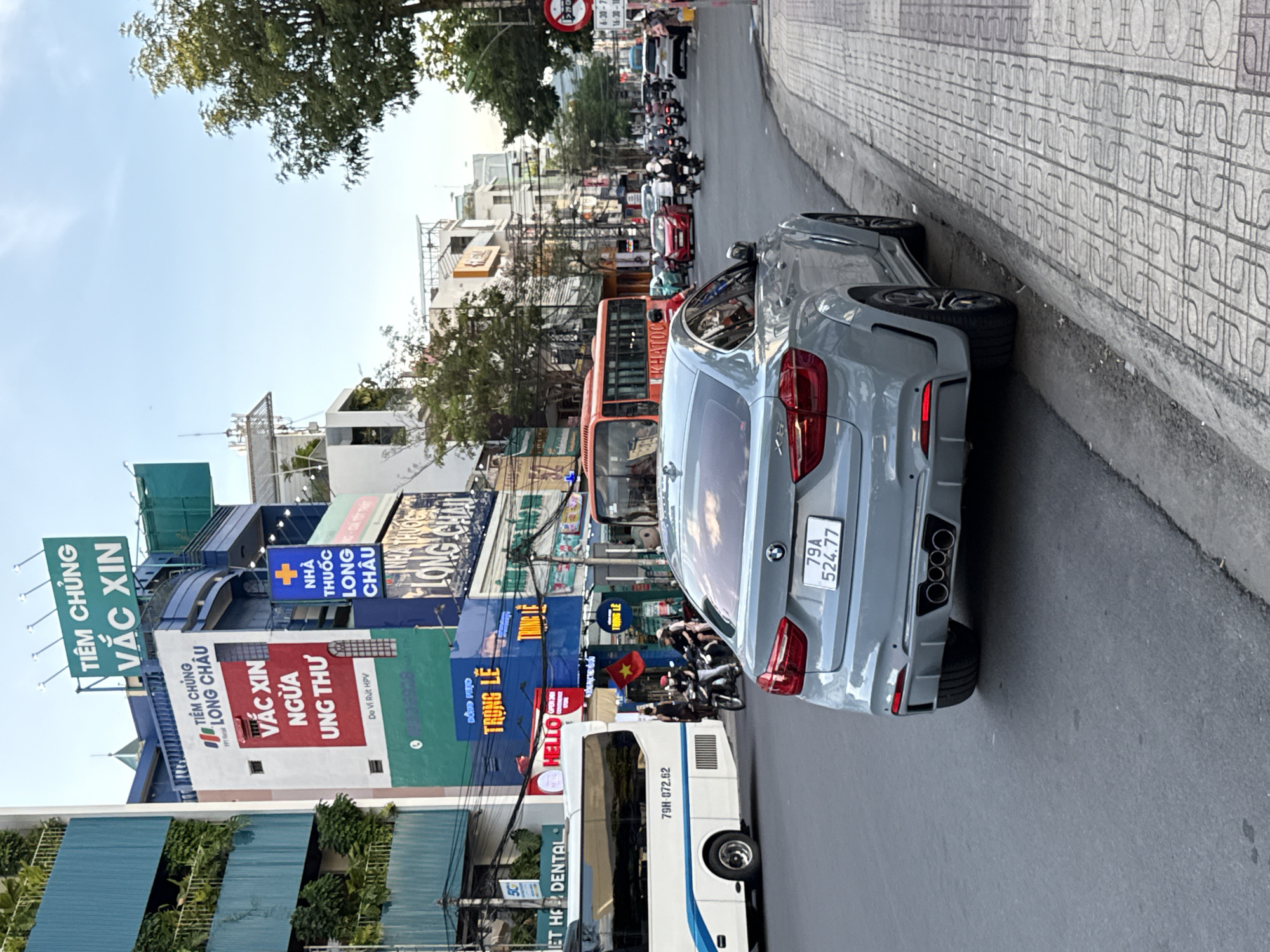 The image depicts a street scene in Nha Trang, Vietnam. In the foreground, a gray BMW X6 is parked on the right side of the road, facing away from the viewer. The car's license plate reads "79A 524.77". The road is paved and has a sidewalk with patterned tiles on the right.
The background is filled with buildings of varying heights and architectural styles, predominantly displaying Vietnamese signage. On the left, a multi-story building is covered in green foliage, with balconies. Several commercial signs are visible, including "TIỆM CHUNG VẮC XIN" (Vaccination Center) and "VẮC XIN NGỪA UNG THƯ" (Vaccine to Prevent Cancer), associated with "LONG CHÂU". Another sign reads "TRONG LÊ".
A white bus with the number "79H 072.62" on its side is traveling on the road, partially obscuring the view of other vehicles and pedestrians. To the right of the bus, several people are seen on motorbikes, and a red bus is visible further down the street. Traffic signals and power lines are present, indicating a busy urban environment. The sky is clear and blue, suggesting daylight.