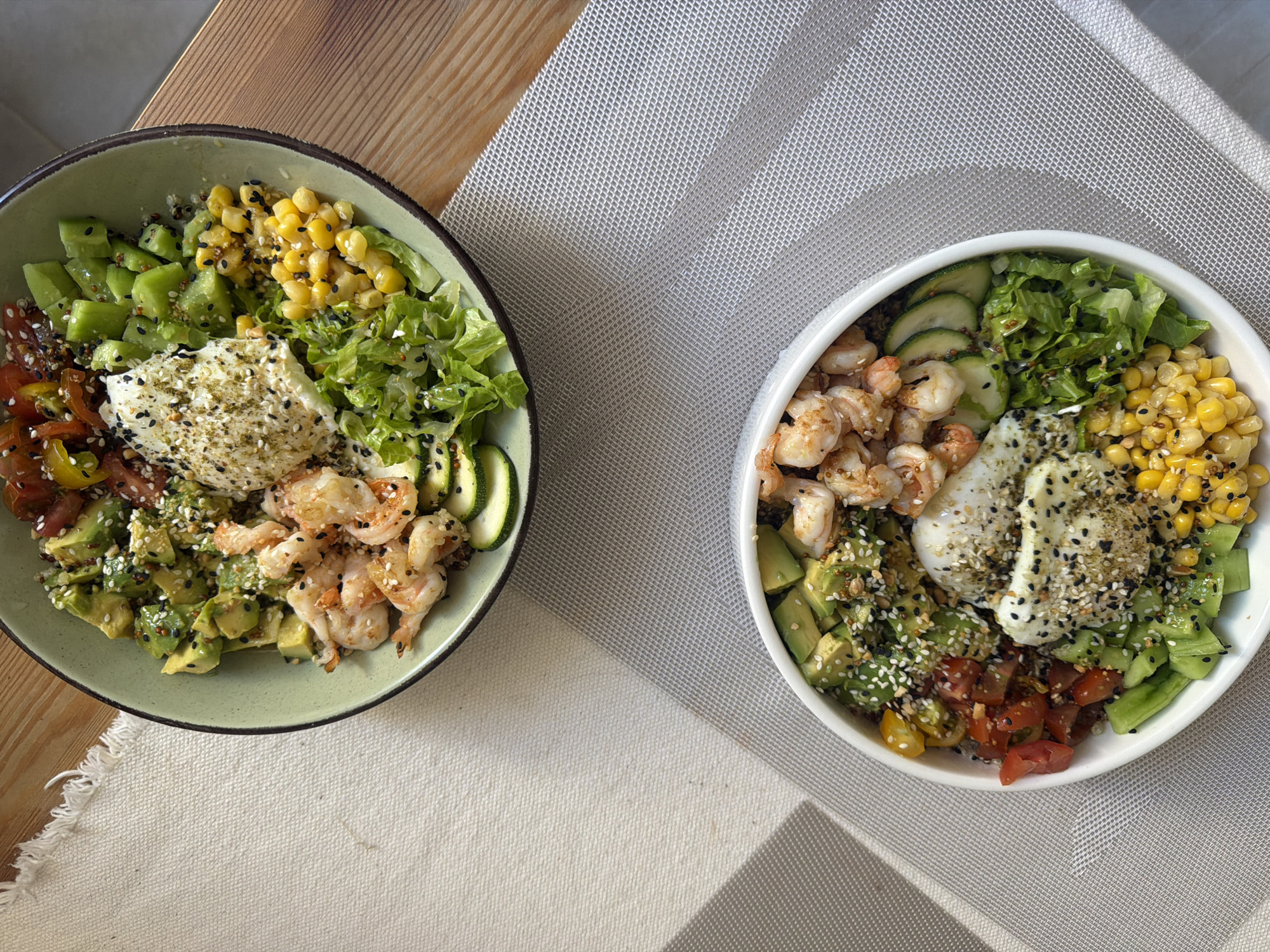 The image displays an overhead view of two individually prepared food bowls, situated in Tel-Aviv, Israel. The upper bowl, light green with a dark rim, rests on a light wood surface partially covered by an off-white, woven fabric placemat with a fringed edge. This bowl contains distinct sections of cooked shrimp, diced avocado, sliced zucchini, green leaf lettuce, corn kernels, diced green vegetables, and diced red and yellow tomatoes. A central dollop of white creamy sauce, topped with black and white sesame seeds and green herbs, is present. The lower bowl is white and placed on a light grey, woven placemat with a grid pattern. Its contents mirror the upper bowl, featuring cooked shrimp, diced avocado, sliced zucchini, green leaf lettuce, corn kernels, diced green vegetables, and diced red and yellow tomatoes. This lower bowl contains two dollops of the same white creamy sauce, similarly garnished with black and white sesame seeds and green herbs. Both bowls show scattered black and white sesame seeds across various ingredients. Natural light entering from the left casts clear shadows on the placemats, indicating an indoor daytime environment. No people are visible.