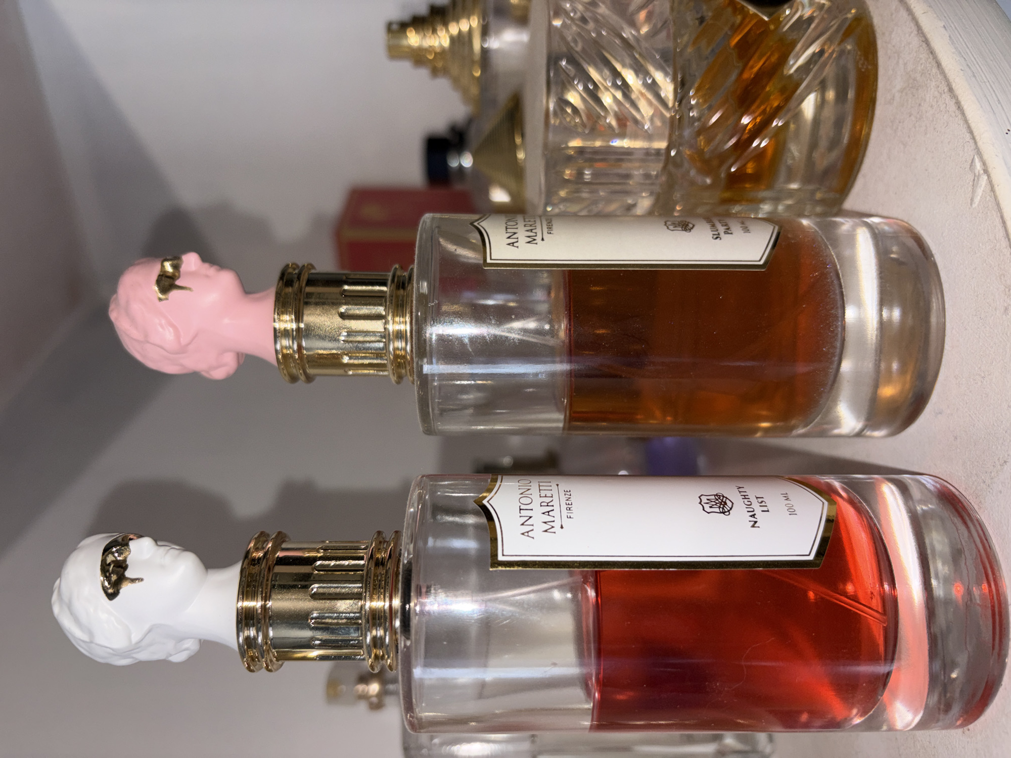 The image depicts two perfume bottles, lying horizontally, stacked on top of each other within a light-colored cabinet or shelf. Both bottles are labeled "Antonio Maretti Firenze." The lower bottle contains a red liquid and is labeled "Naughty List" and "100 ml." The upper bottle contains a brown liquid and is labeled "Slutty Party" and "100 ml." Each bottle is topped with a gold-colored sprayer and a bust of a head; one head is white and the other is pink. Other perfume bottles of various shapes and styles are partially visible in the background. No people are visible. The location is unknown; there is no indication that it is Izhevsk, Russia.
