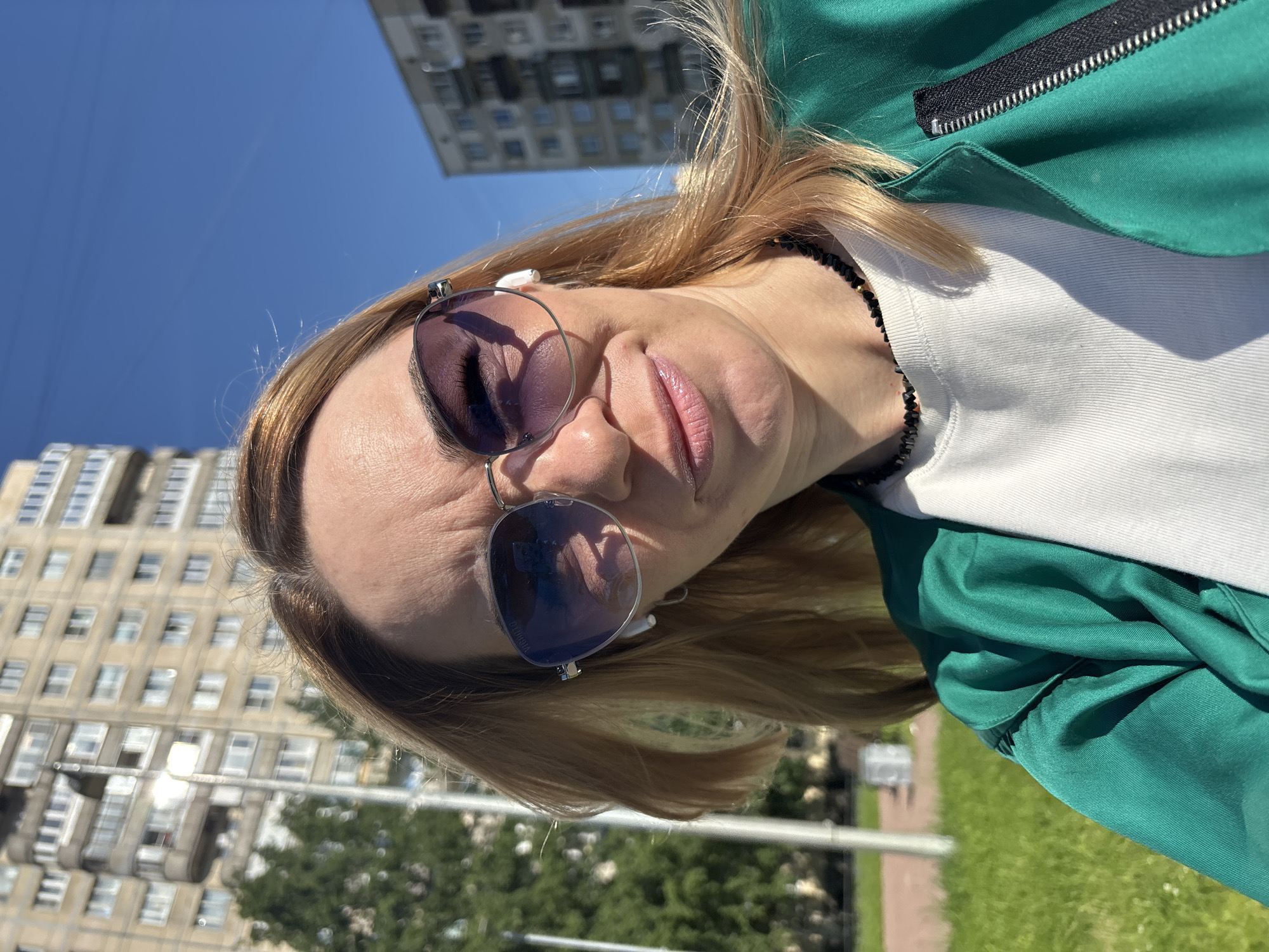 A close-up, low-angle shot features a woman outdoors on a clear, sunny day. Her head is tilted slightly upwards, and she appears to be looking towards the camera. She has light brown or blonde hair, styled around her face. She wears large, aviator-style sunglasses with purple-tinted lenses; the brand name "VIRROVA" is faintly visible on the left lens. A white wireless earbud is placed in her left ear. Her lips are slightly parted in a neutral expression.
She is dressed in a bright green jacket with a visible silver zipper, worn over a white t-shirt. A black beaded necklace with small golden accents is visible around her neck.
The background consists of a clear, cloudless blue sky, indicating bright, sunny weather, likely midday or early afternoon. Several multi-story residential buildings, common in urban settings, are visible in the distance. Green trees and a patch of green grass are also present, along with what appears to be a paved path or road. The setting suggests a casual moment in an urban park or street area.