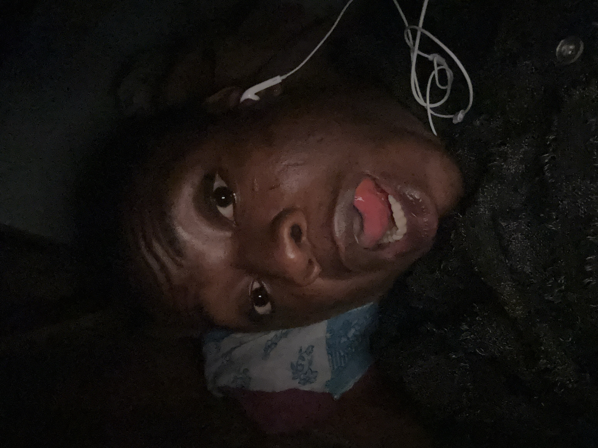 A close-up, low-light image shows a person lying on their back, with their head resting on a patterned pillow. The person has dark skin, and their mouth is open, revealing their tongue and teeth. They are wearing white earphones. The location is Unknown, Unknown.