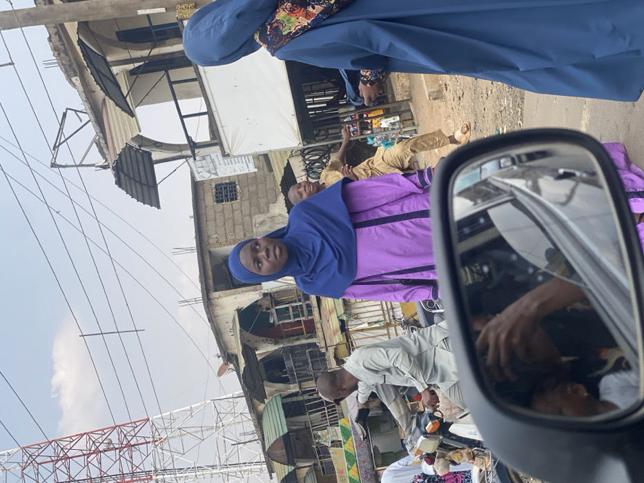 An eye-level shot shows several people on a street with aged buildings and overhead power lines. A woman in a purple hijab and dress stands in the foreground, looking directly at the camera. A partially visible rearview mirror reflects the interior of a car. An individual in blue hijab and dress stands in the background. Other people are scattered throughout the scene. The buildings appear to be shops or businesses.