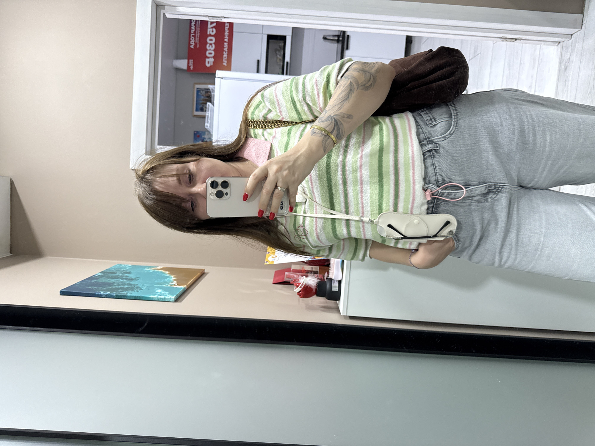 A woman with brown hair and bangs takes a selfie in a mirror. She is wearing a green and white striped collared shirt, light gray jeans, and a brown fuzzy purse over her shoulder. She holds a white phone with a white phone case attached to a lanyard with a pink drawstring. Her right hand is on her hip, and her left hand holds the phone. A tattoo is visible on her left arm. Behind her, a white cabinet holds several items, including a red rose in a clear vase, a bottle, and a comb. A framed picture of a beach is hanging on the wall to the left. In the background, a doorway opens to a room with a white refrigerator and kitchen cabinets. A red sign with Cyrillic lettering is visible on the refrigerator. The floor is tiled with a light gray pattern. The environment appears to be an indoor residential setting, possibly an apartment in Sochi, Russia.