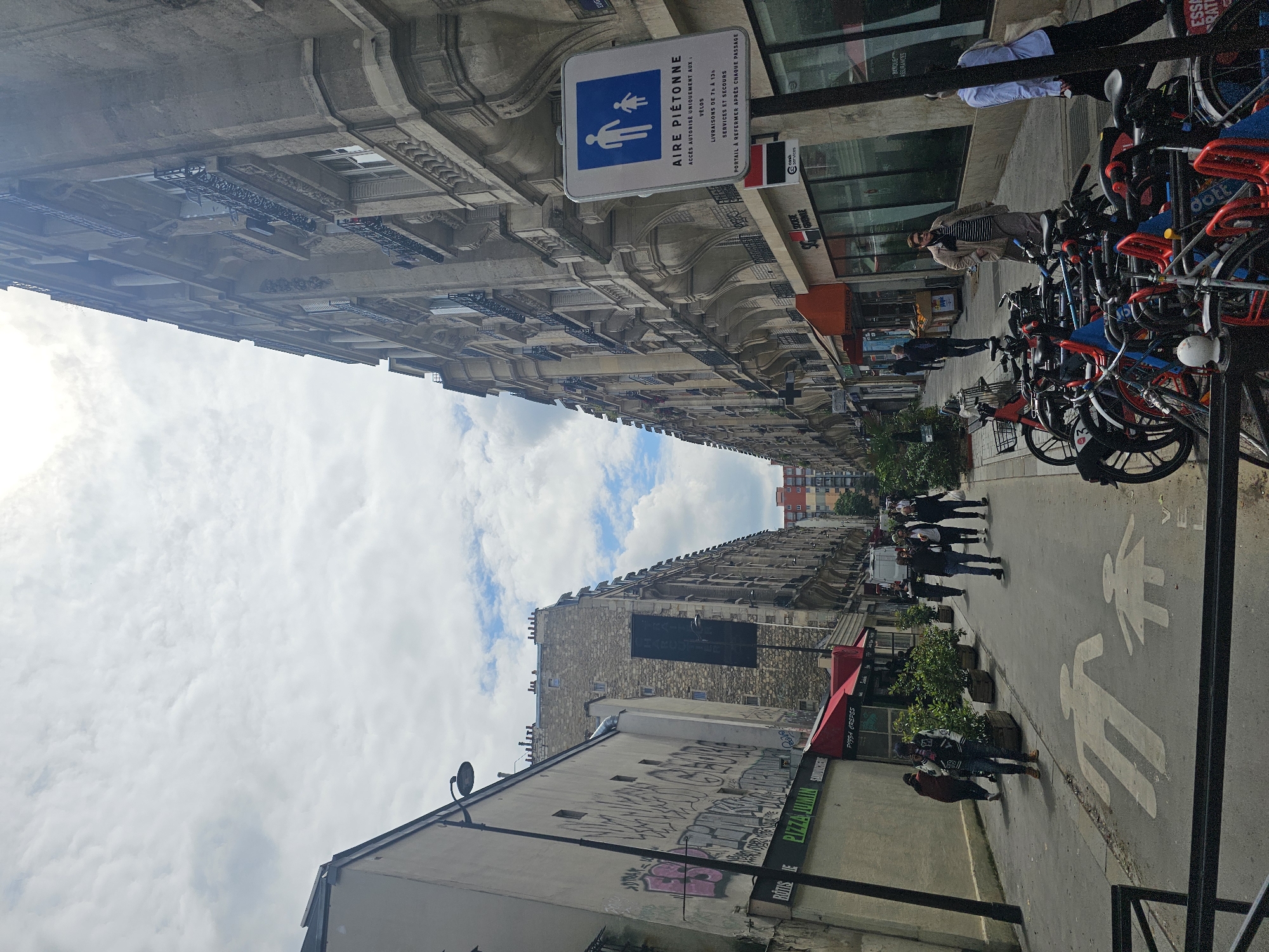 The image shows a street in Paris, France, designated as a pedestrian zone ("Aire Piétonne"), as indicated by the street sign. People are walking along the street, which also has markings on the pavement indicating a shared pedestrian and bicycle space. Bicycles are parked along the side of the street. The buildings on either side of the street are tall, classic Parisian structures with detailed facades. A pizza shop is visible on one side of the street with graffiti on the wall adjacent to it. The sky is partly cloudy.