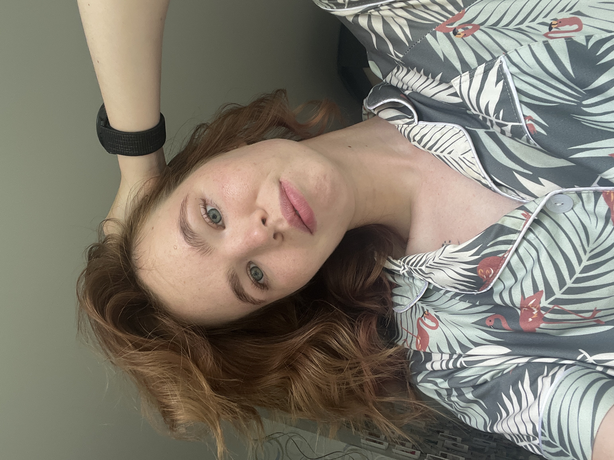A young woman with auburn hair and blue eyes is shown in a close-up, selfie-style shot. She is wearing a pajama top with a flamingo and palm leaf print. Her right arm is raised, with her hand resting on her head, and she is wearing a black fitness tracker on her wrist. The background is plain and out of focus, with a hint of a window or light source to the left. No specific location in Chaikovsky, Russia, is identifiable from the image.