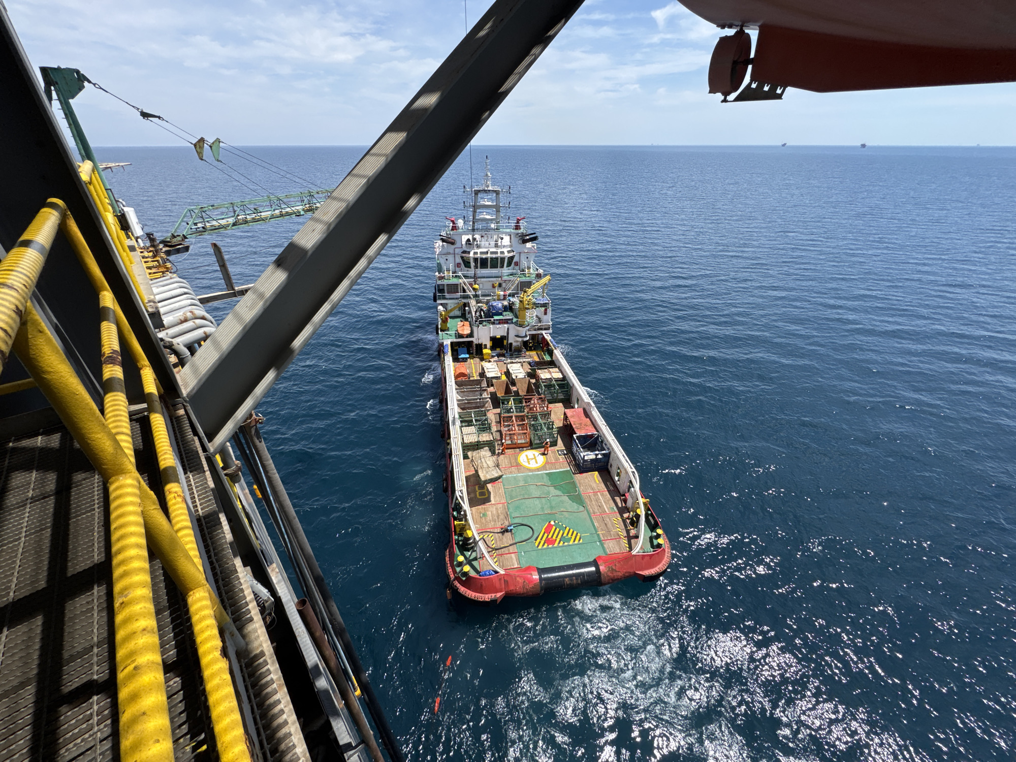 An offshore supply vessel is docked alongside a larger structure, possibly an oil rig or another ship, in a bright, daytime ocean setting. The larger structure occupies the left foreground, with its grey metal beams and yellow-striped pipes visible. A green crane extends from this structure towards the vessel. The supply vessel, painted red and white, has a helideck marked with a white 'H' on a blue circle, and its deck is laden with cargo, including metal cages and pipes. The sea is a deep blue, with a clear sky overhead dotted with white clouds. Several other ships are visible in the distance on the horizon. The scene appears to be in an industrial offshore environment.