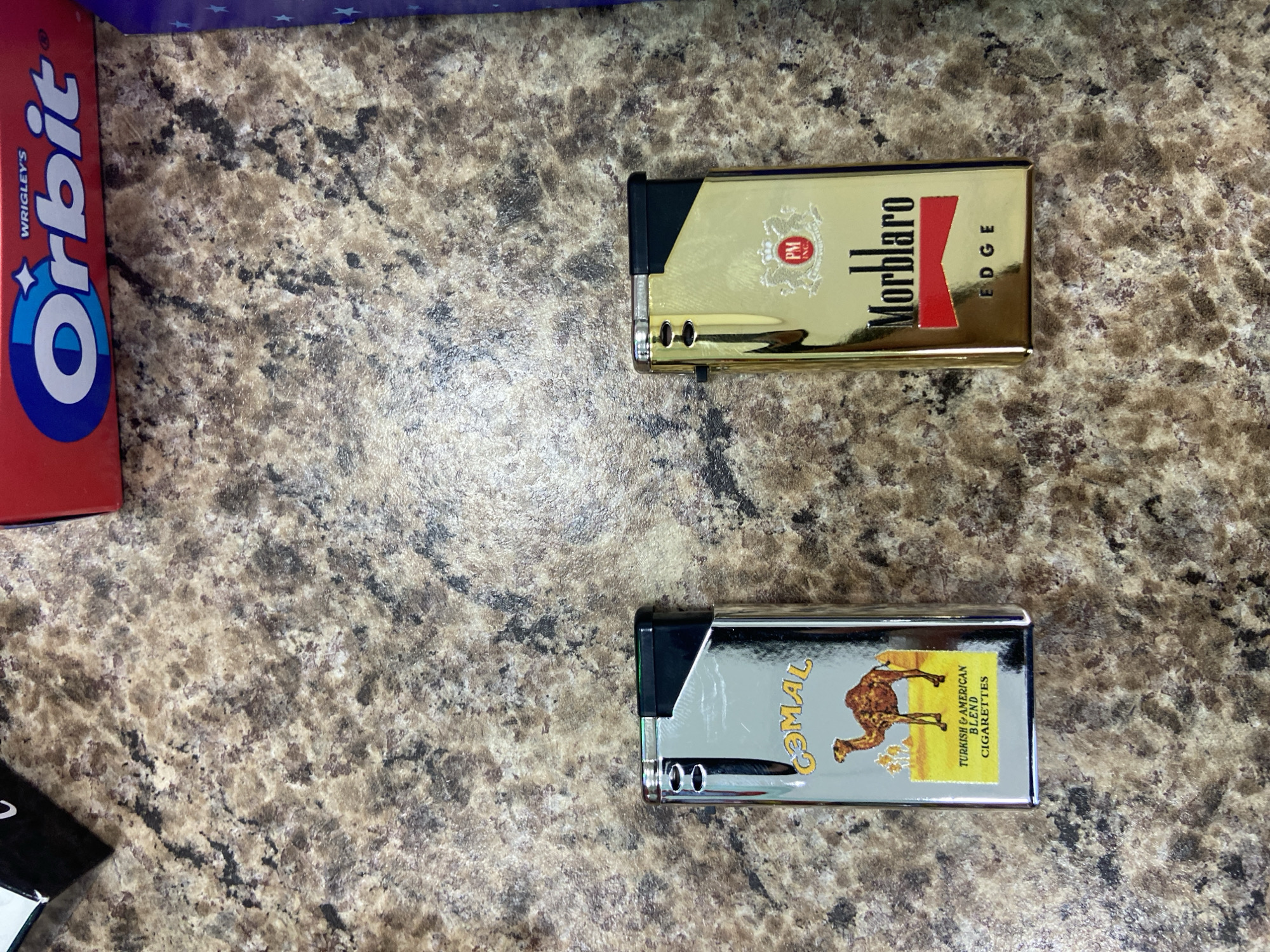This is an overhead shot of two cigarette lighters and a box of Orbit gum placed on a speckled, marble-like countertop. The subjects are the lighters and gum, with the countertop serving as the setting.
The top lighter is gold with a black accent and features the branding "Morblaro EDGE" in red and black text. A circular logo with "PM INC." is also visible on the lighter.
Below the gold lighter is a silver lighter with a black accent. It displays the "Camel" brand logo, which includes a camel silhouette and text that reads "TURKISH & AMERICAN BLEND CIGARETTES."
To the left of the lighters is a red and blue box of "Orbit" gum, with the brand name prominently displayed in white and blue lettering.
There are no people visible, and no specific activity or event is depicted beyond the arrangement of the objects. The lighting suggests an indoor setting, likely during the daytime, with no indication of weather conditions or specific time of day. The location context of Yegorlykskaya, Russia, is provided, but there are no visual cues in the image to confirm this.