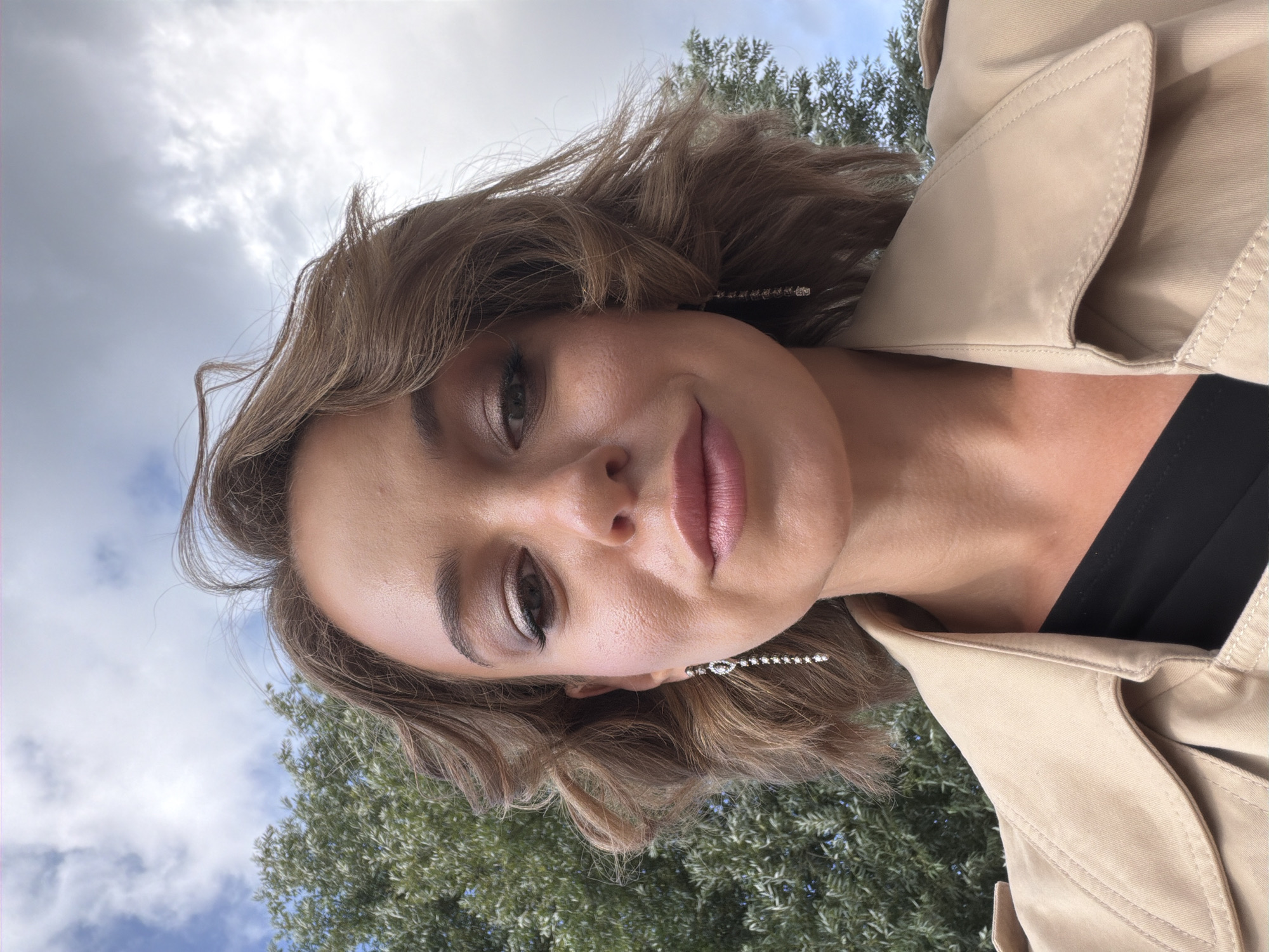 A woman is taking a selfie outdoors with a cloudy sky and trees in the background. She has short, wavy brown hair, and is wearing a light beige trench coat over a black top. She has on makeup and long, dangly earrings. The lighting suggests it is daytime.