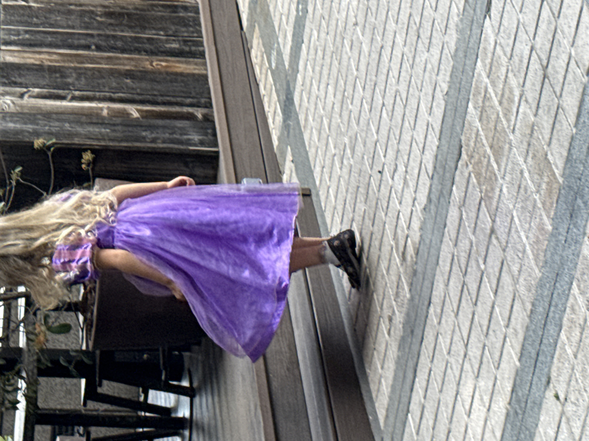 A girl in a purple dress is lying on a patterned concrete surface next to a wooden railing, with some of the scene oriented at an angle. In the background, there's a metal staircase and some greenery. It is impossible to know if this image was captured in the city of Barcelona, Spain, or not.