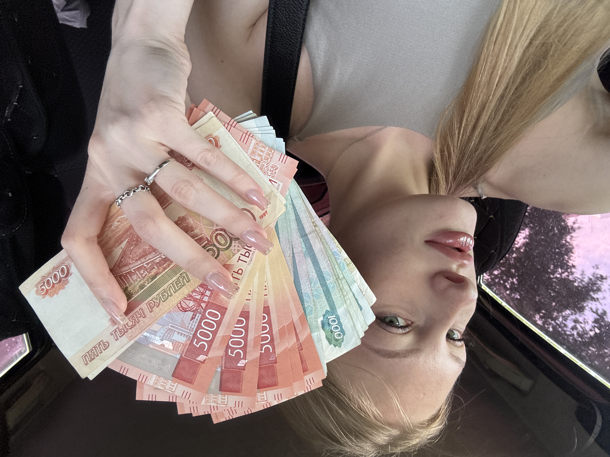 A young woman with blonde hair and green eyes is sitting in a car, holding a fan of Russian Ruble banknotes. She is wearing a gray tank top and a silver ring on her left hand. The banknotes are in denominations of 1000 and 5000 rubles. The background shows the interior of the car and a blurred view of trees outside the window, with hints of purple and pink colors, possibly indicating sunset or sunrise. The image does not contain specific landmarks that would allow for precise location identification within the city of Perm, Russia.