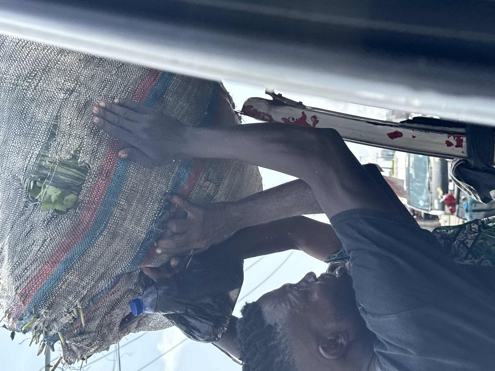 A man secures a burlap sack to the roof rack of a vehicle in Lagos, Nigeria. He is dark-skinned, wearing a dark T-shirt, and has closely cropped hair. He is reaching up with both arms, his hands pressing against the sack to hold it in place. The burlap sack is held in place by thin bungee cords connected to the roof rack. The sack is striped with red and blue and contains green leafy material. A blue plastic water bottle and black plastic bag dangle from the sack. The vehicle's roof rack is visible, and the exterior environment suggests a bustling urban setting.