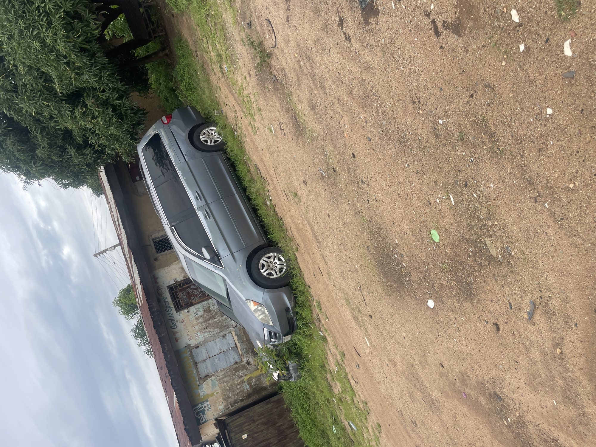 A grey minivan is parked on a dirt road in front of a dilapidated building in New Stadium, Nigeria. The building has a corrugated metal roof and its walls are covered in peeling paint and graffiti. The minivan is facing right, and its front is partially obscured by overgrown green bushes. The ground is covered in sand and small stones, with scattered litter including plastic bottles and wrappers. A large tree with dense green foliage stands to the right of the minivan, casting a shadow on the ground. The sky is overcast with grey clouds. There are no people visible in the image.