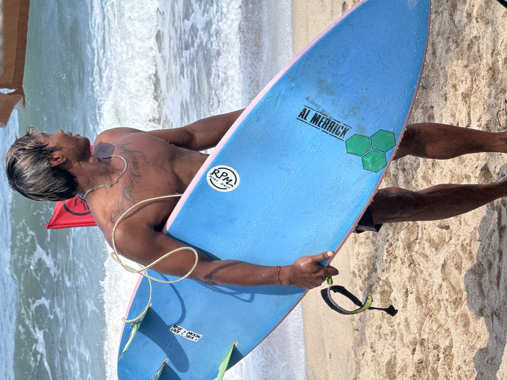 A surfer, with sandy blond hair and a lean build, stands on a beach in Kuta, Indonesia, holding a bright blue surfboard with pink rails. He is shirtless, revealing tattoos on his chest and shoulder. The surfboard has several stickers and the brand "AL MERRICK" is visible. The ocean waves are in the background, with a red flag planted in the sand. The sand is a light brown, with footprints and shadows indicating a sunny day.