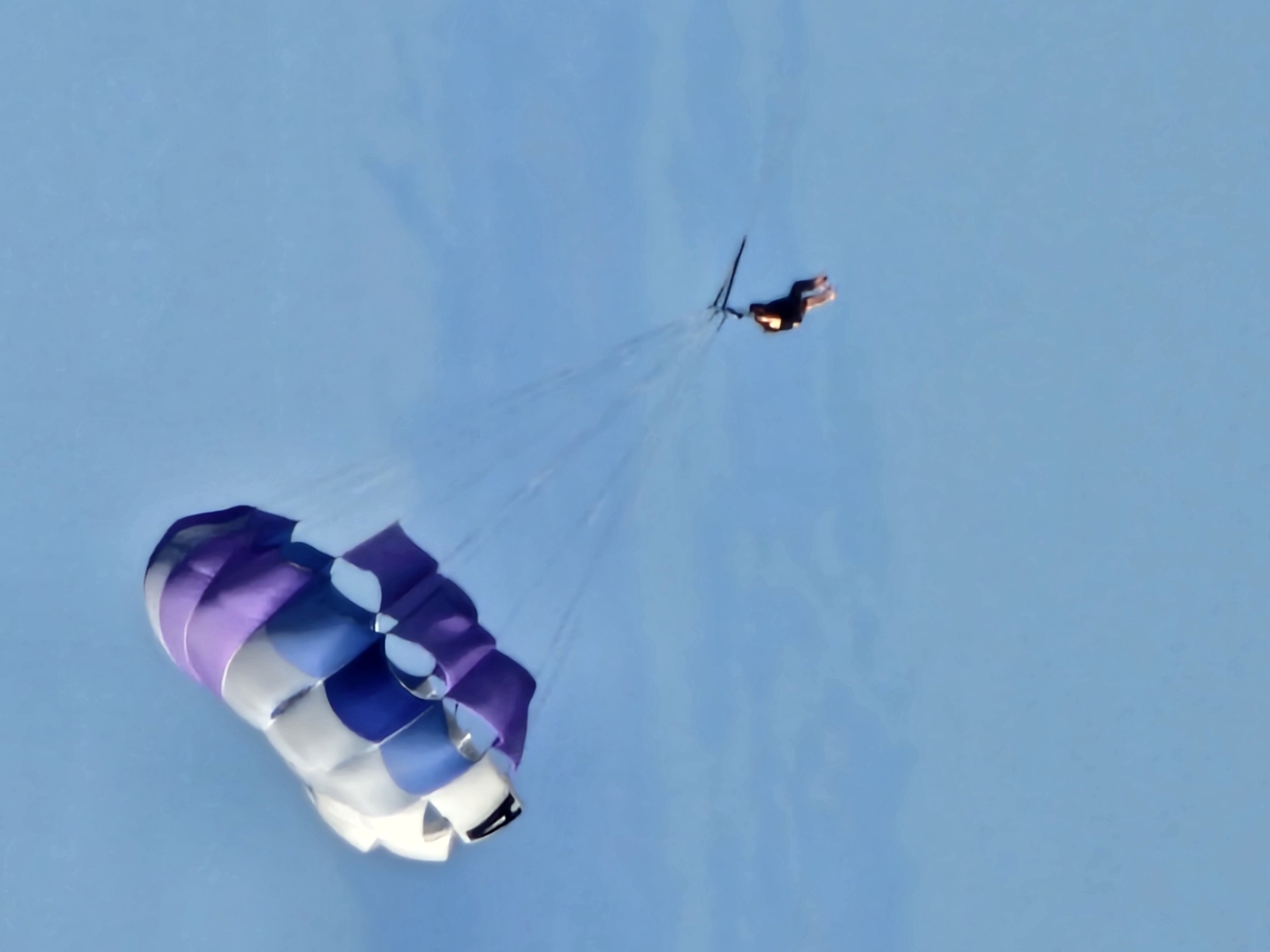 A person is parasailing in the sky, attached to a parachute that is patterned with purple, blue, and white panels. The background is a clear blue sky. The location appears to be Kuta Selatan, Indonesia.