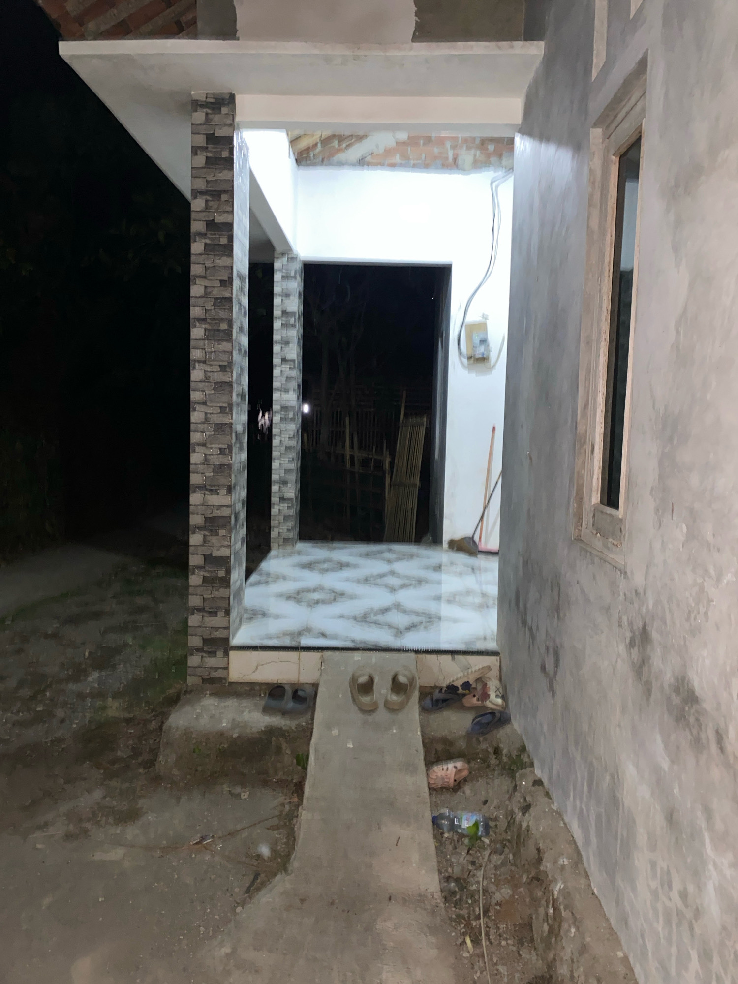 The image depicts an outdoor scene at night, likely in a residential area of Planjan, Indonesia. In the foreground, a concrete pathway leads towards the entrance of a building. Several pairs of sandals and flip-flops are scattered on and beside the pathway, indicating recent use. A trench or shallow excavation is visible to the right of the pathway, containing some debris, including a plastic bottle and a wire.
The building's entrance is framed by pillars, some of which are covered in textured stone tiles, while others appear to be unfinished concrete or plaster. The floor inside the entrance is tiled with a geometric pattern. Beyond the entrance, a doorway leads into a darker interior space. A broom with a wooden handle leans against a wall near the doorway, and electrical wiring is visible on the exterior wall. To the left of the entrance, a dark, overgrown area suggests the presence of vegetation. The overall impression is of an unfinished or newly constructed area with a utilitarian and somewhat disheveled appearance. The lighting suggests it is nighttime or a very dimly lit area.