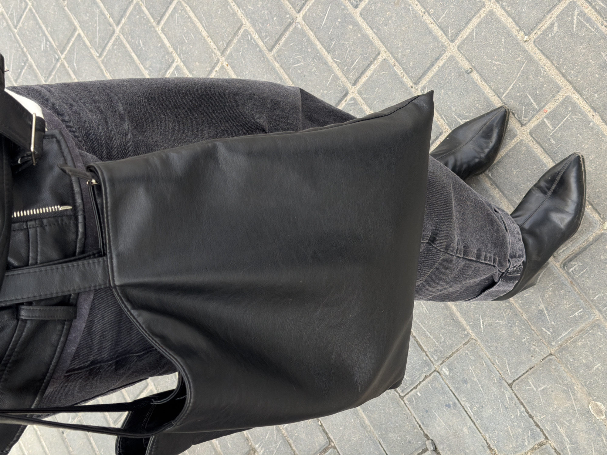 An overhead shot depicts a person's legs and torso. They wear dark gray jeans and black pointed-toe ankle boots. A black leather jacket is partly visible, with a silver zipper detail. A large black leather bag obscures much of the view. The background shows gray paving stones. The location is Засечное, Russia.
