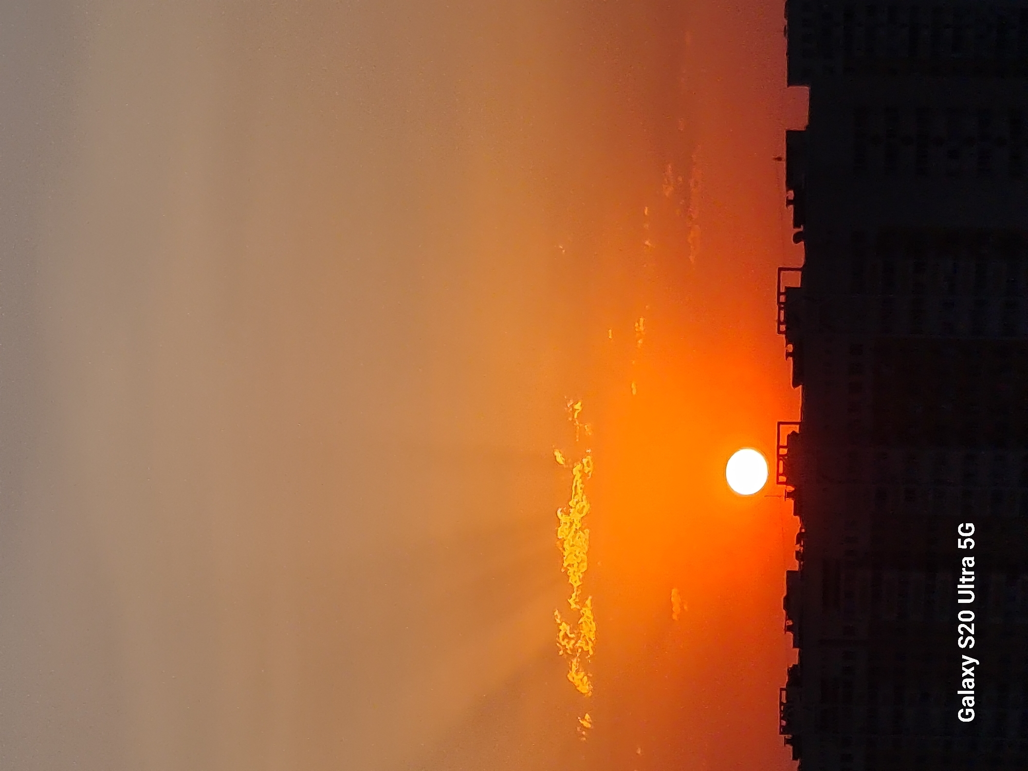 This image shows a sunset over the city of Almaty, Kazakhstan. The sun is a bright orange circle low on the horizon, partially obscured by the silhouetted rooftops of apartment buildings. Rays of sunlight fan out from the sun through scattered clouds. The sky is a gradient of orange and beige, with darker shades near the horizon. The foreground is dominated by the dark silhouettes of buildings. The text "Galaxy S20 Ultra 5G" is visible in the bottom left corner of the image.