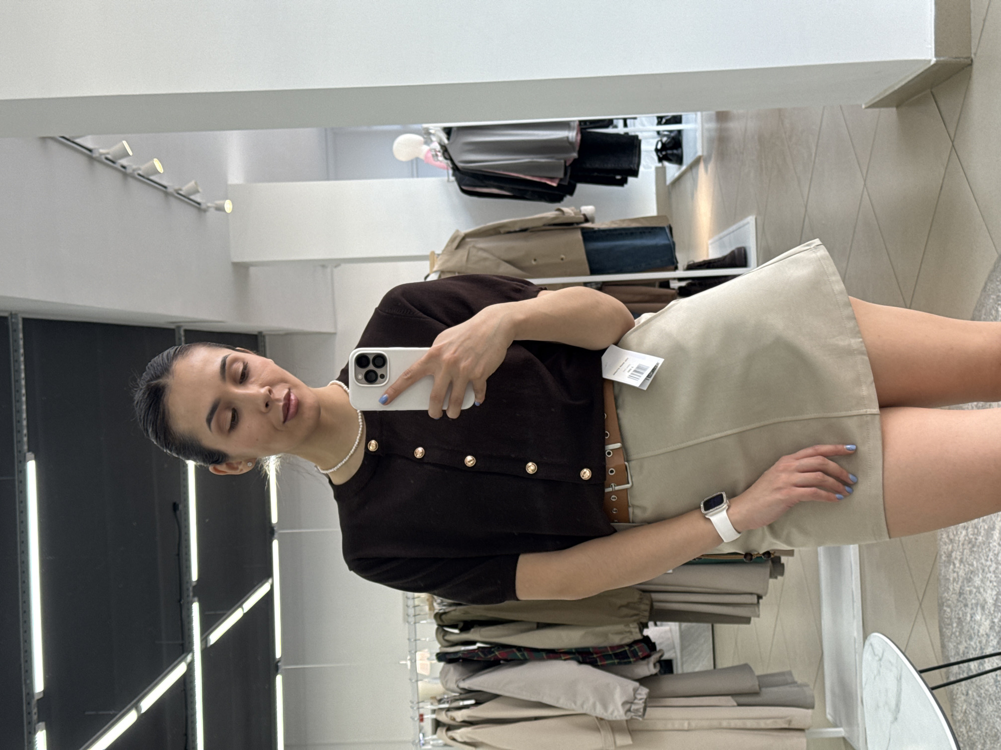 A woman is taking a mirror selfie in a clothing store. She is wearing a brown, short-sleeved cardigan with gold buttons, a beige mini skirt with a brown belt, and a pearl necklace. She is holding a white smartphone. The background shows racks of clothing. The location is not specified.