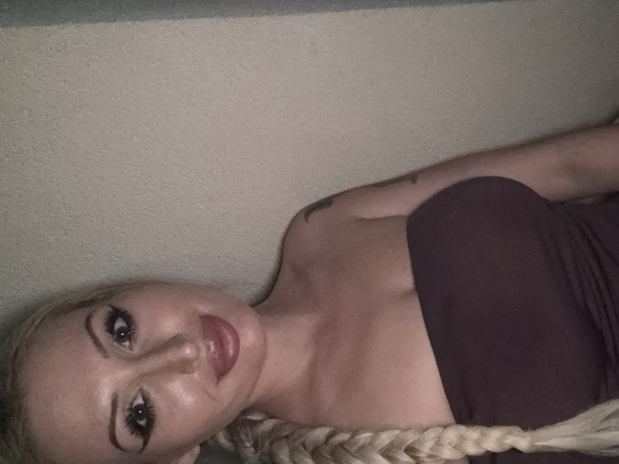 A blonde woman with heavy makeup, including winged eyeliner and plumped lips, is shown from the chest up in a close-up portrait. She has a long braid of blonde hair that falls over her left shoulder. She is wearing a strapless dark maroon top. A tattoo is visible on her upper right arm. The background consists of a textured, light-colored wall. The lighting is dim, creating shadows.