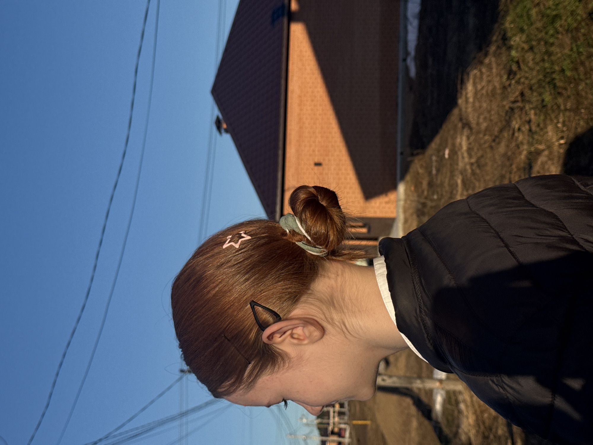 A medium, eye-level shot depicts a woman with auburn hair pulled into a bun, secured with a star-shaped hairpin and a hair tie. She is wearing a black puffer jacket over a white collared shirt. Her face is in profile, turned towards the right. Behind her, out of focus, is a red brick building with a gable roof casting a sharp shadow, a strip of dirt and grass, and a clear blue sky with power lines. Based on visual cues, the image likely originates from Chaikovsky, Russia.