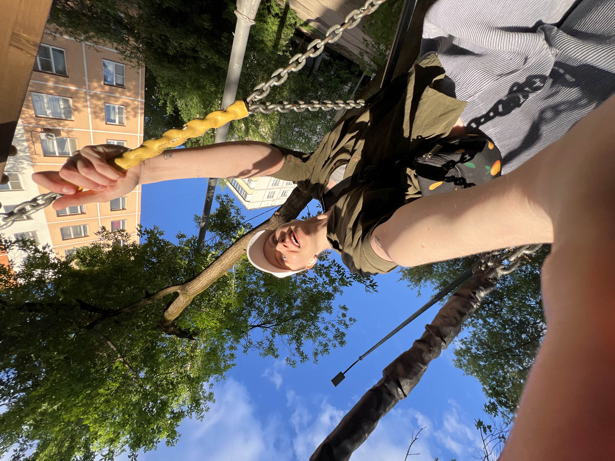 A low-angle shot shows a person holding onto a yellow bar suspended by chains. The person is wearing a white cap, green shirt, and striped pants with a small black bag. Trees with green leaves fill the background, along with a blue sky dotted with clouds. A building with orange exterior walls and windows is visible in the upper left corner. The location is Khimki, Russia.