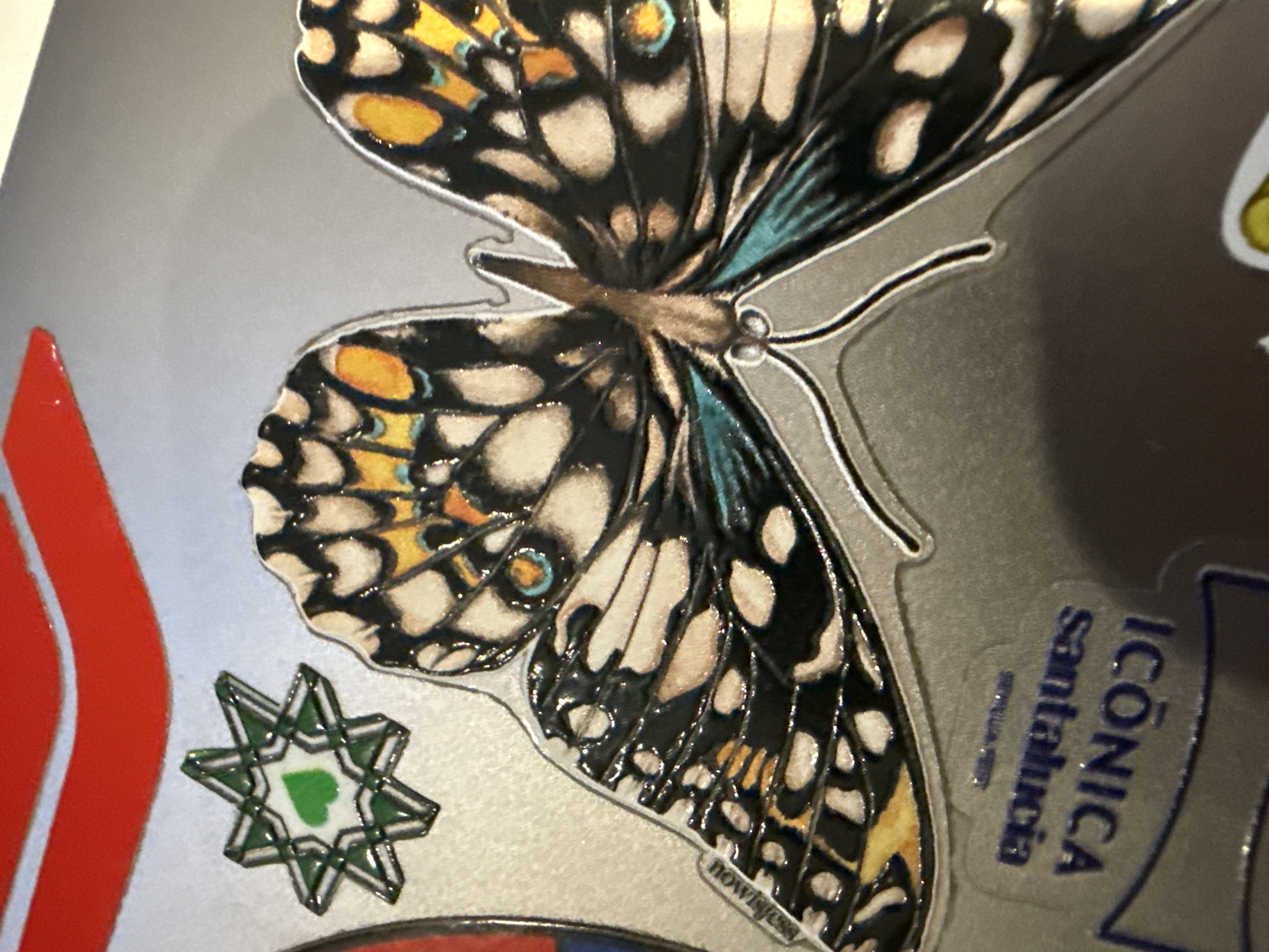 A close-up shot showcases various stickers on a silver surface. Predominantly featured is a butterfly sticker with black, white, yellow, and teal accents. To the left, there's a red sticker and a star-shaped sticker with a green heart at its center. In the bottom right corner, a blue sticker reads "ICÓNICA santa lucía." A portion of another sticker is also visible. The stickers are likely located in Santa Lucía, a neighborhood in Mexico City, Mexico, suggested by the “ICÓNICA santa lucía” sticker.