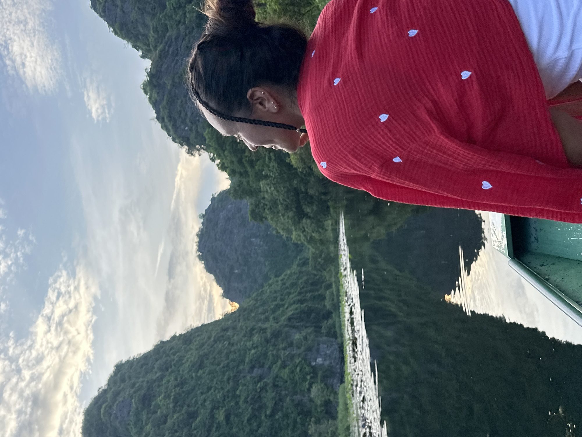 The image depicts a woman in the foreground, viewed from the back and right side, looking up and to the left. She has her hair pulled up in a bun with a braid accent. She is wearing a red, textured top adorned with small white hearts and a white garment underneath. The woman is seated in a green boat, which is partially visible in the bottom right corner.
The background features a scenic natural landscape characterized by lush green, karst mountains rising steeply from a calm body of water. The water's surface reflects the sky and the surrounding mountains, with a scattering of lily pads or aquatic plants on the left side. The sky is partly cloudy, with soft sunlight breaking through, suggesting either sunrise or sunset. The overall setting is tranquil and picturesque, indicative of a location like the Trang An or Tam Coc area in Nam Hoa Lư Ward, Vietnam, known for its similar karst formations and waterways.