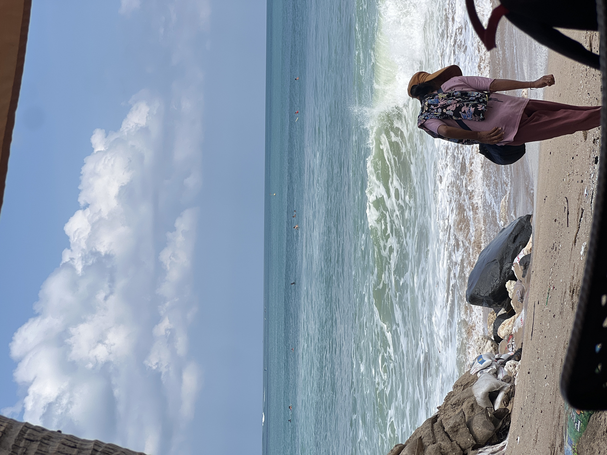A person wearing a pink shirt, pink pants, a floral scarf, and a brown hat stands on a sandy beach in Kuta, Indonesia, facing away from the camera and towards the ocean. The person is carrying a black bag over their shoulder. The ocean is a turquoise blue with white waves crashing on the shore. Several surfers can be seen in the distance. Large rocks and scattered trash are visible on the beach. The sky is blue with scattered white clouds. A palm tree trunk is visible on the left side of the image.
