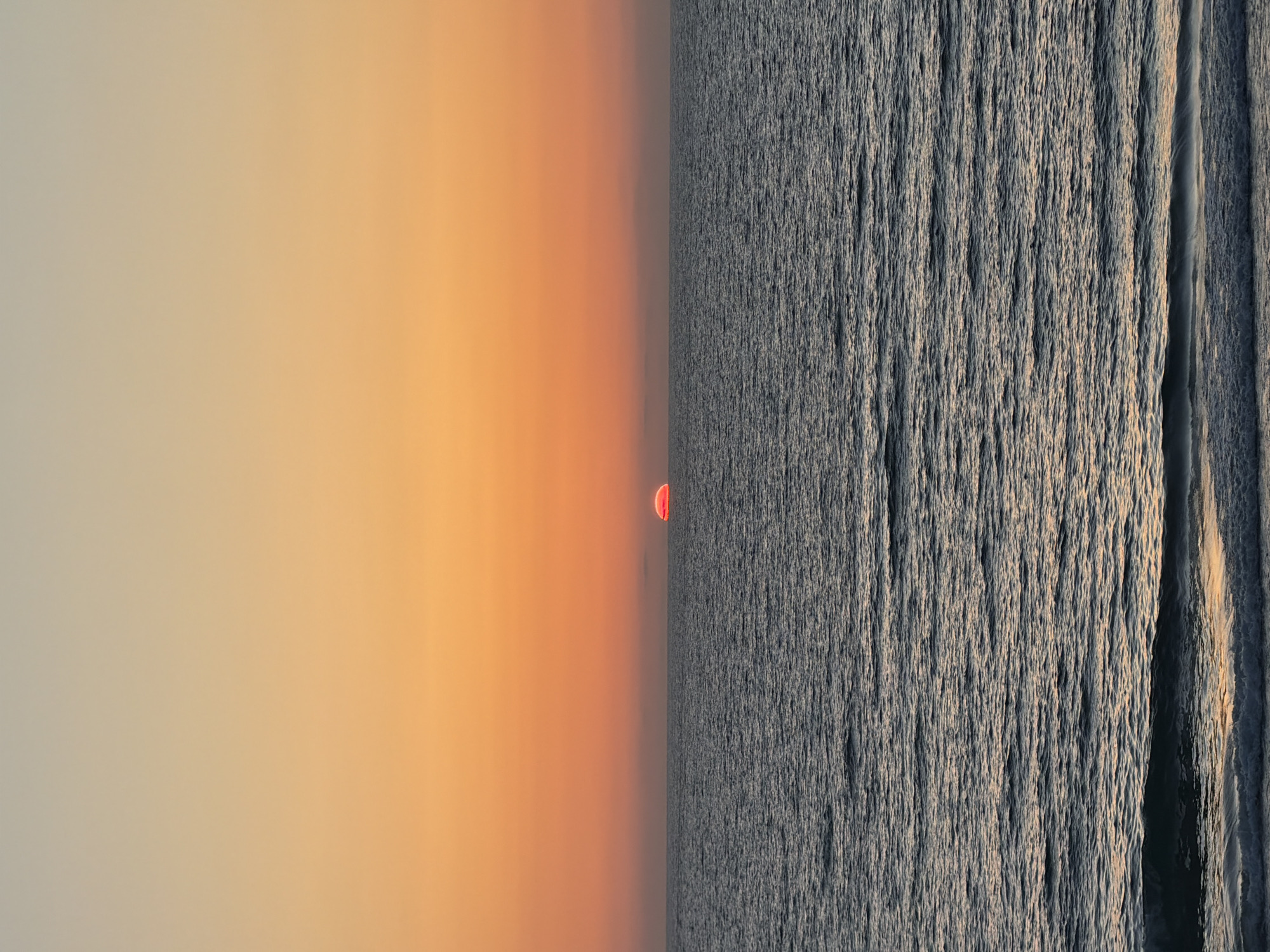 The image depicts a sunset over the Mediterranean Sea, taken from Tel-Aviv, Israel. The left side of the image showcases a gradient of colors, transitioning from a pale yellow to a deep orange near the horizon. A portion of the red sun is visible on the horizon line, appearing partially submerged, while the other side is choppy water. The texture of the water creates a contrast with the smooth gradient of the sky. A vertical line, potentially a wake, cuts through the water.