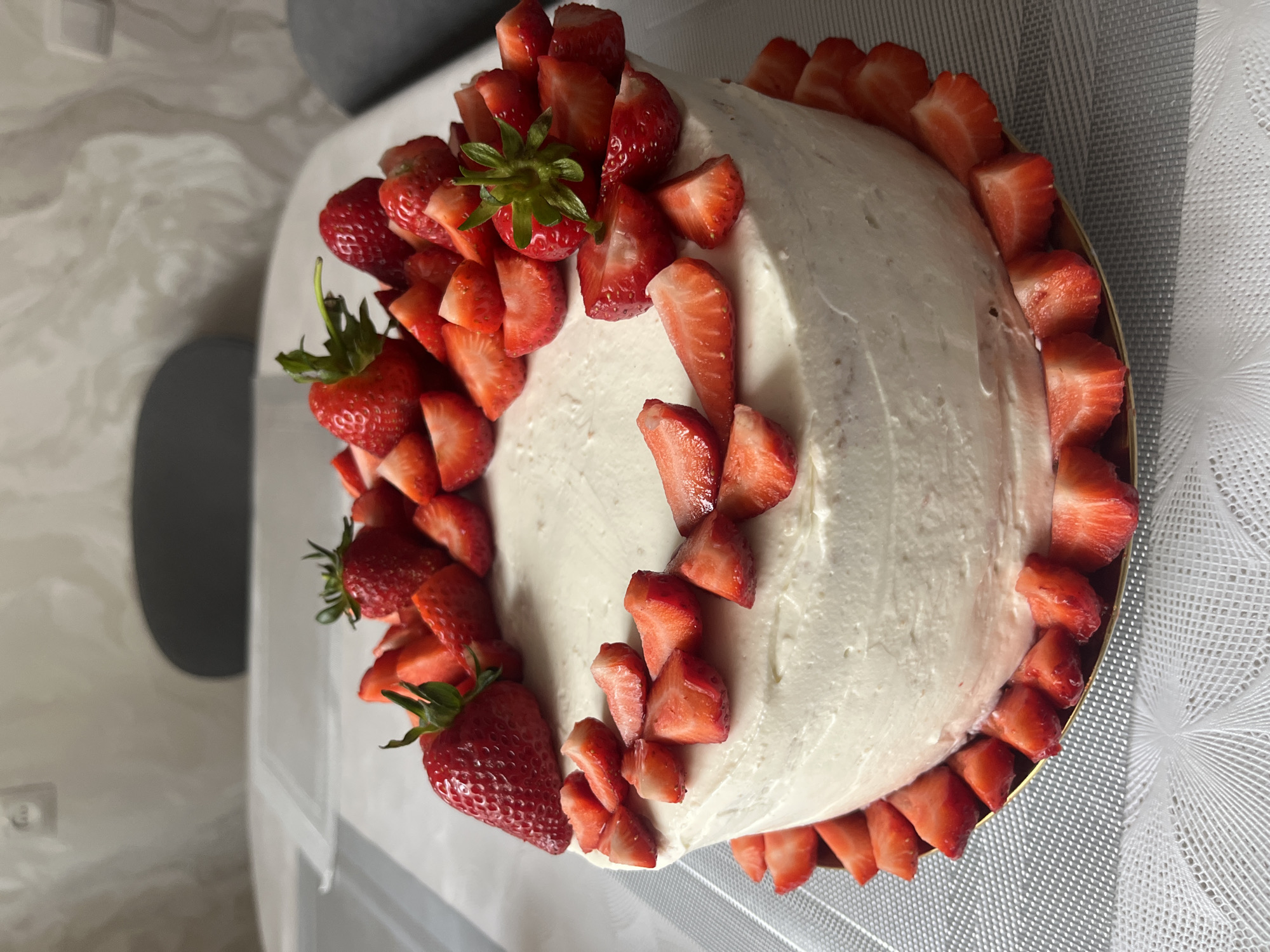 The image shows a strawberry cake, topped with white frosting and decorated with fresh strawberries. Some strawberries are whole with green leaves, while others are sliced. The cake sits on a gold-colored plate on a table covered with a white, patterned tablecloth. The background is blurry, but a gray surface is visible. The image does not provide information about the location, people, actions, or interactions. It does not indicate that it was taken in Kaspiysk, Russia.