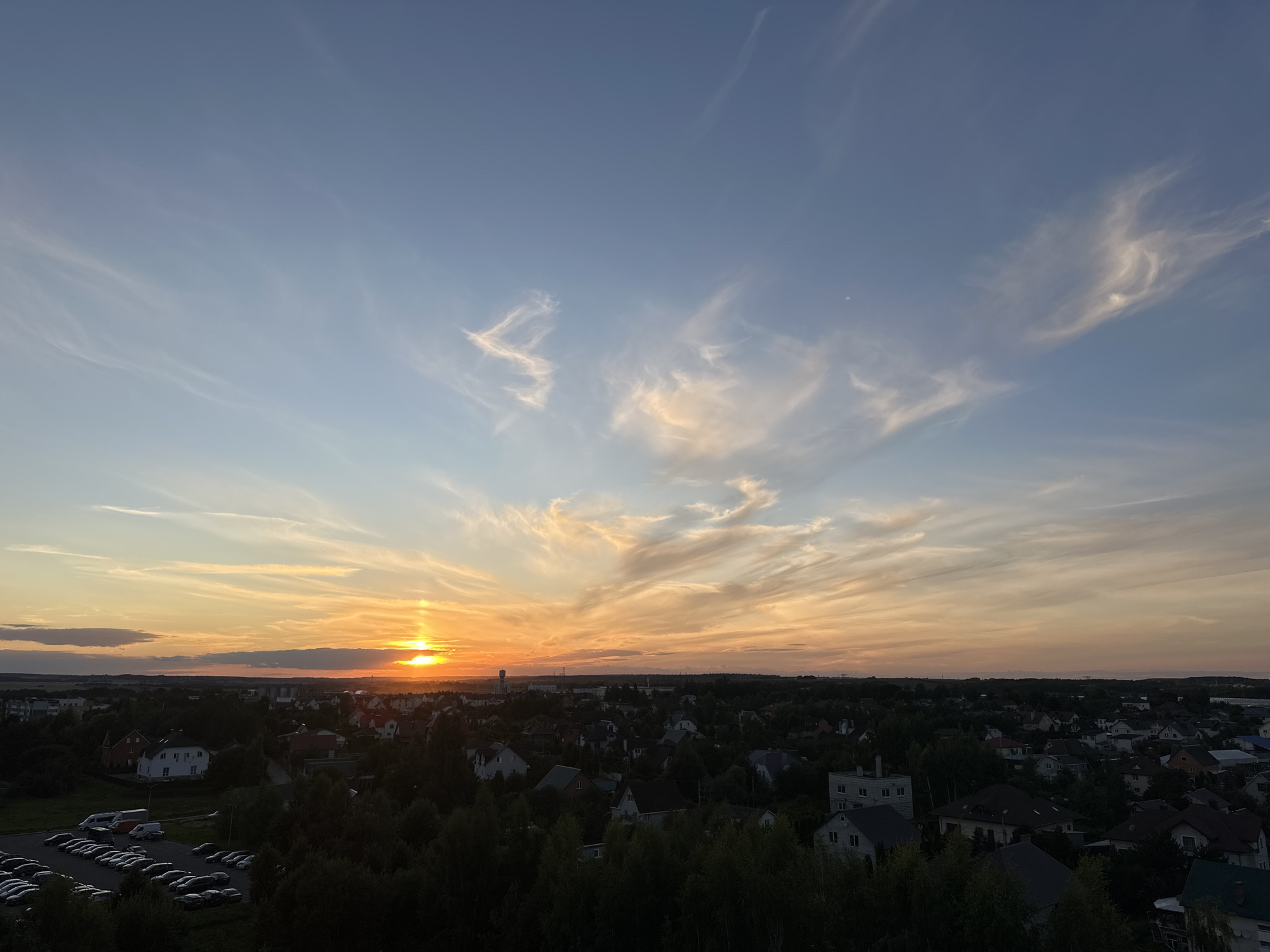 The image depicts an aerial view of a suburban landscape in Shchomyslitski Sel'ski Sovet, Belarus, during a sunset. The sky is a gradient of blues and oranges, with wispy clouds illuminated by the setting sun, which is visible on the left horizon. The foreground is dominated by a dense canopy of dark green trees. Below the trees lies a residential area with numerous houses, varying in architectural style and roof color, interspersed with patches of vegetation. A parking lot filled with cars is visible in the lower left quadrant of the image. In the distance, a water tower stands as a prominent structure among a scattering of buildings. The overall scene captures a tranquil evening in a rural or semi-rural Belarusian settlement.