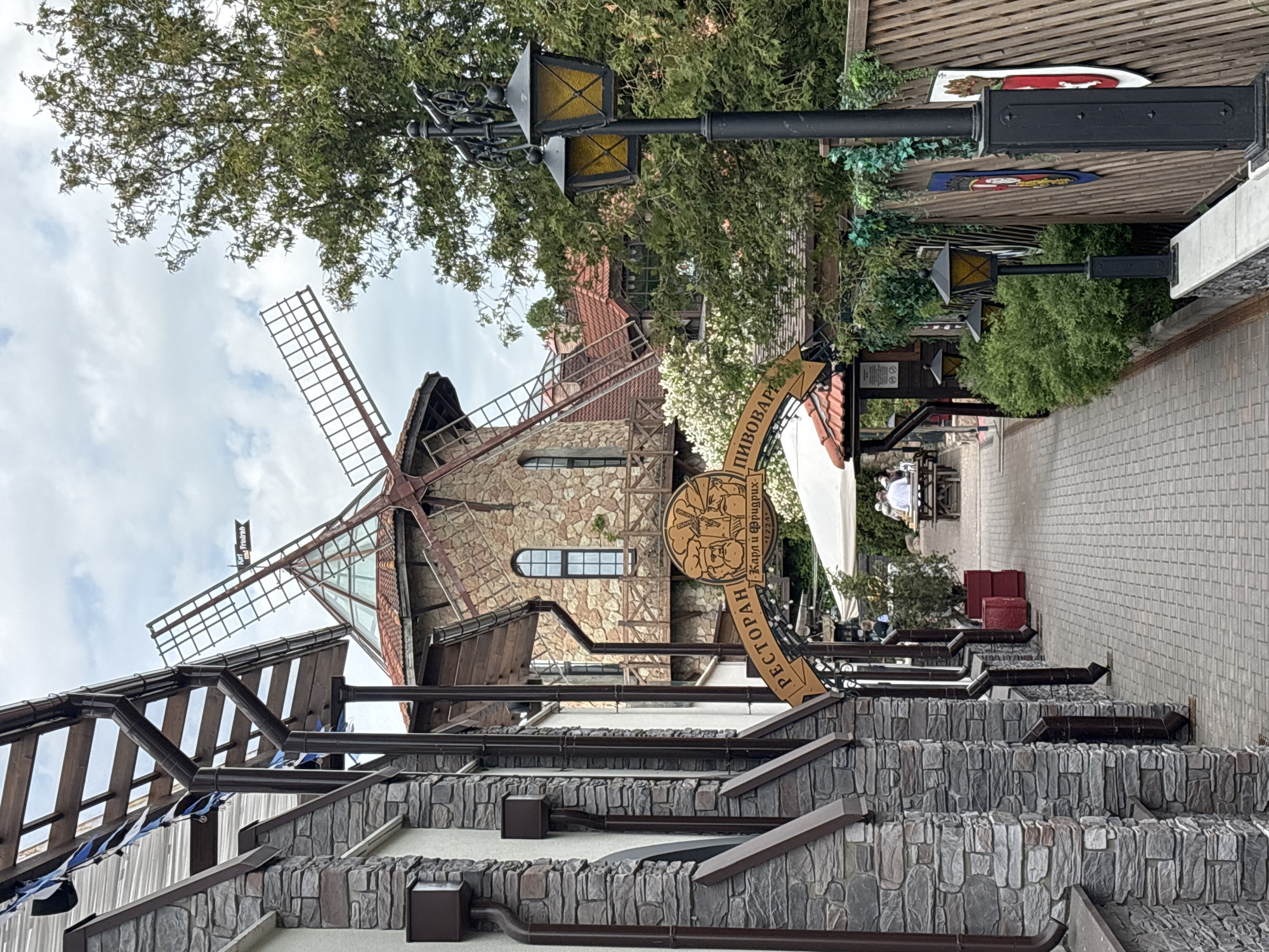 This outdoor scene captures a multi-level restaurant or brewery complex, likely in Saint Petersburg, Russia, featuring a prominent building designed to resemble a stone windmill. The sky above is partly cloudy, suggesting daytime.
The central structure is a two-story stone building with a reddish-brown tiled roof and large windows, topped with a large, four-bladed windmill sail. One of the sails has a small black banner with "Karl Friedrich" written on it. In front of this building, a significant wooden sign, shaped like a beer barrel or coaster, hangs from an ornate pole. The sign is emblazoned with the Cyrillic text "ПИВОВАРНЯ" (Pivovarnya - Brewery) and "РЕСТОРАН" (Restoran - Restaurant). Below the barrel, it reads "КАРЛ И ФРИДРИХЪ" (Karl i Fridrikh - Karl and Friedrich) and the year "1724", suggesting a historical or German theme for the establishment.
The foreground features stacked stone walls, forming terraced levels, with dark brown gutter-like structures and pipes running along them. Small blue and white decorative banners are visible strung along a railing on one of these levels. Ornate black lampposts with yellow-tinted lanterns are positioned throughout the area, surrounded by lush green trees and bushes. A paved pathway, made of light-colored square pavers, extends towards the right side of the image.
In the background, an outdoor dining area is visible under white awnings or umbrellas, where several people are seated at tables, enjoying the setting. Red elements, possibly planters or bins, are also scattered in this area. The overall atmosphere appears tranquil and inviting, typical of a leisure or dining venue.