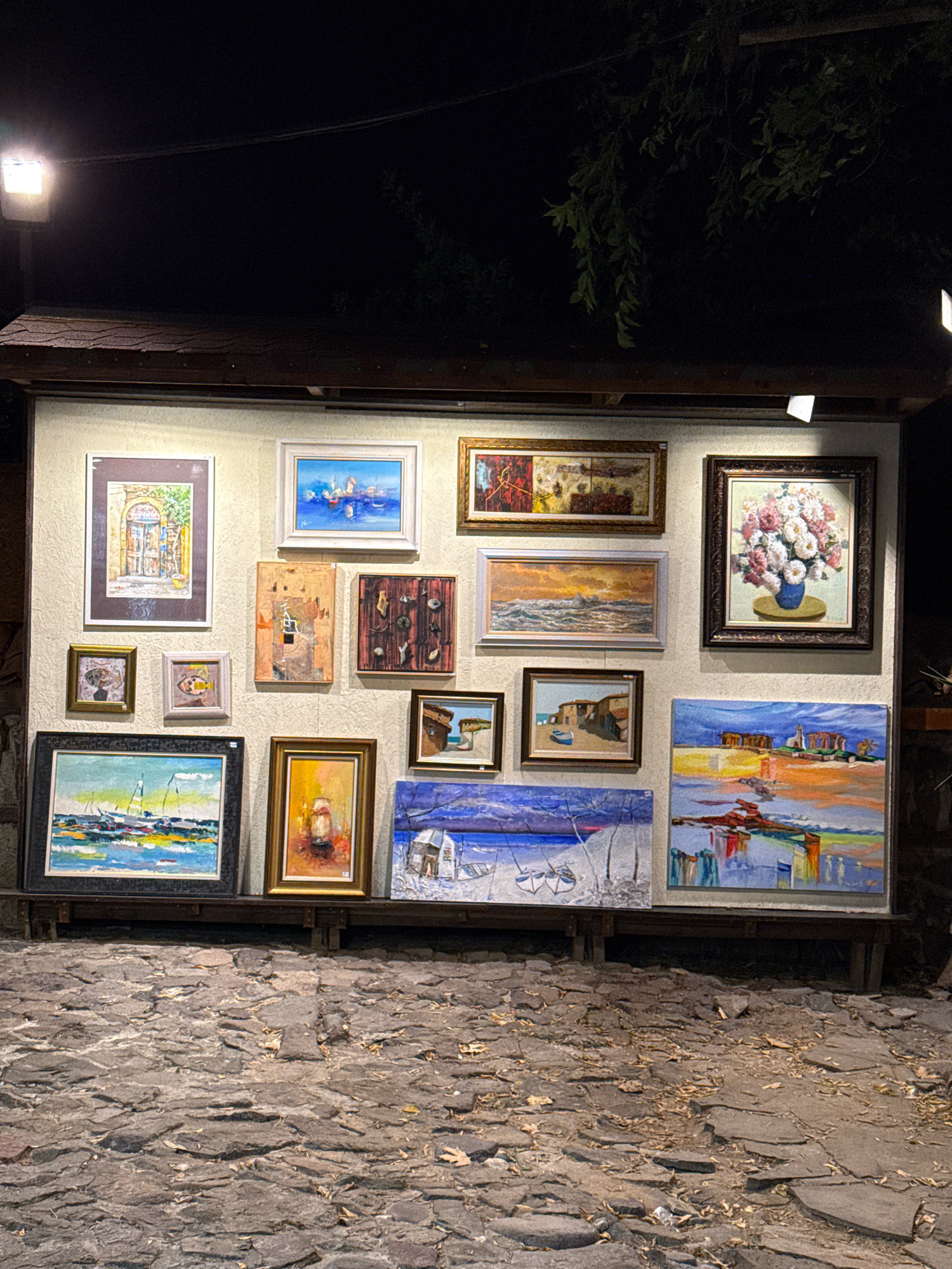 An outdoor art exhibition is displayed at night on a cobbled street in Sozopol, Bulgaria. A light fixture above illuminates the artwork, which consists of several paintings in various sizes and frames, hung on a textured wall. The paintings depict landscapes, seascapes, still life, and architectural scenes. In the foreground, the uneven, gray stone pavement is visible, with scattered dry leaves and debris. The overall scene suggests a quiet evening in a historic town setting.