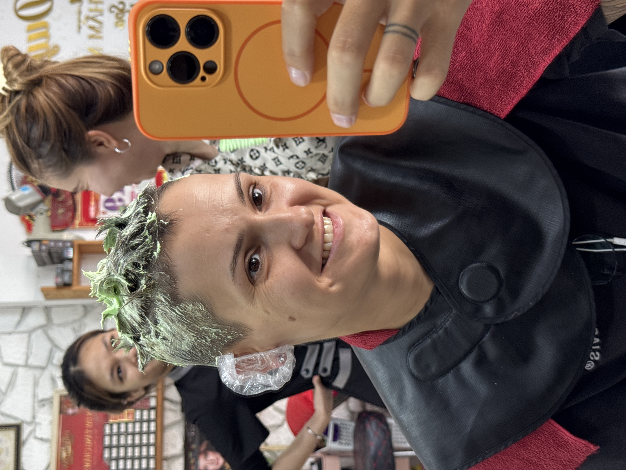 In a hair salon in Nha Trang Ward, Vietnam, a woman with short, dyed green hair is taking a selfie with her phone, which has an orange case. She is smiling, and plastic coverings are on her ears. She is wearing a black smock with a red towel draped around her shoulders. In the background, another woman is blurred, with her hair tied up in a bun. The salon appears to be well-lit, with shelves of hair products visible on the wall.