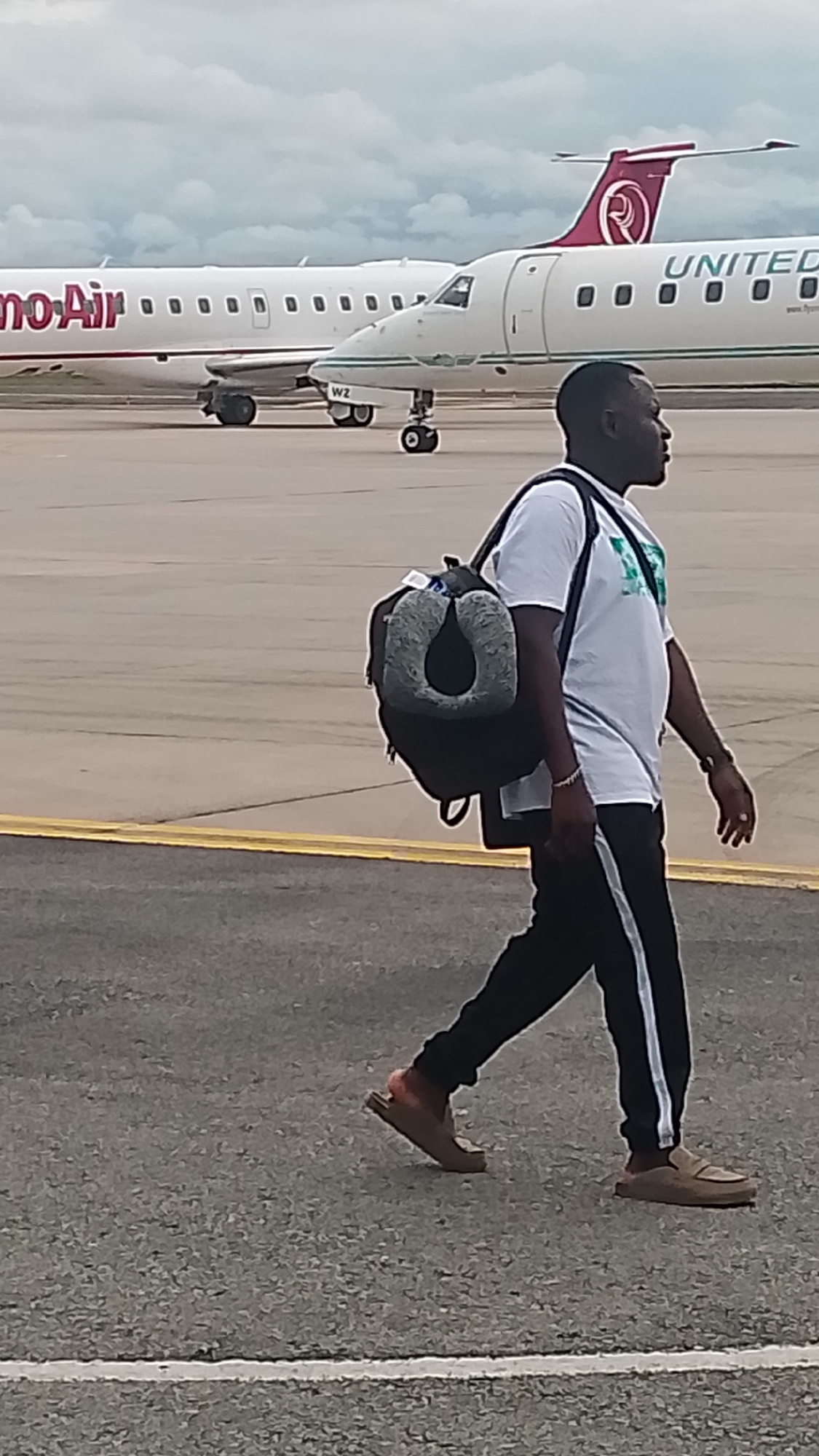 A medium shot captures a man in profile walking on an airport tarmac in Orerokpe, Nigeria. He is wearing a white t-shirt with a green design, black pants with a white stripe, and slip-on shoes. A dark backpack with a gray neck pillow is slung over his left shoulder. The man appears to be walking away from the camera. In the background, two commercial airplanes are parked on the tarmac under a cloudy sky. The airplane on the left has "mo Air" in pink lettering on its fuselage. The airplane on the right is identified as "UNITED" in large green lettering, with a burgundy and gold logo on its tail. The tarmac is paved with asphalt and marked with yellow and white lines.