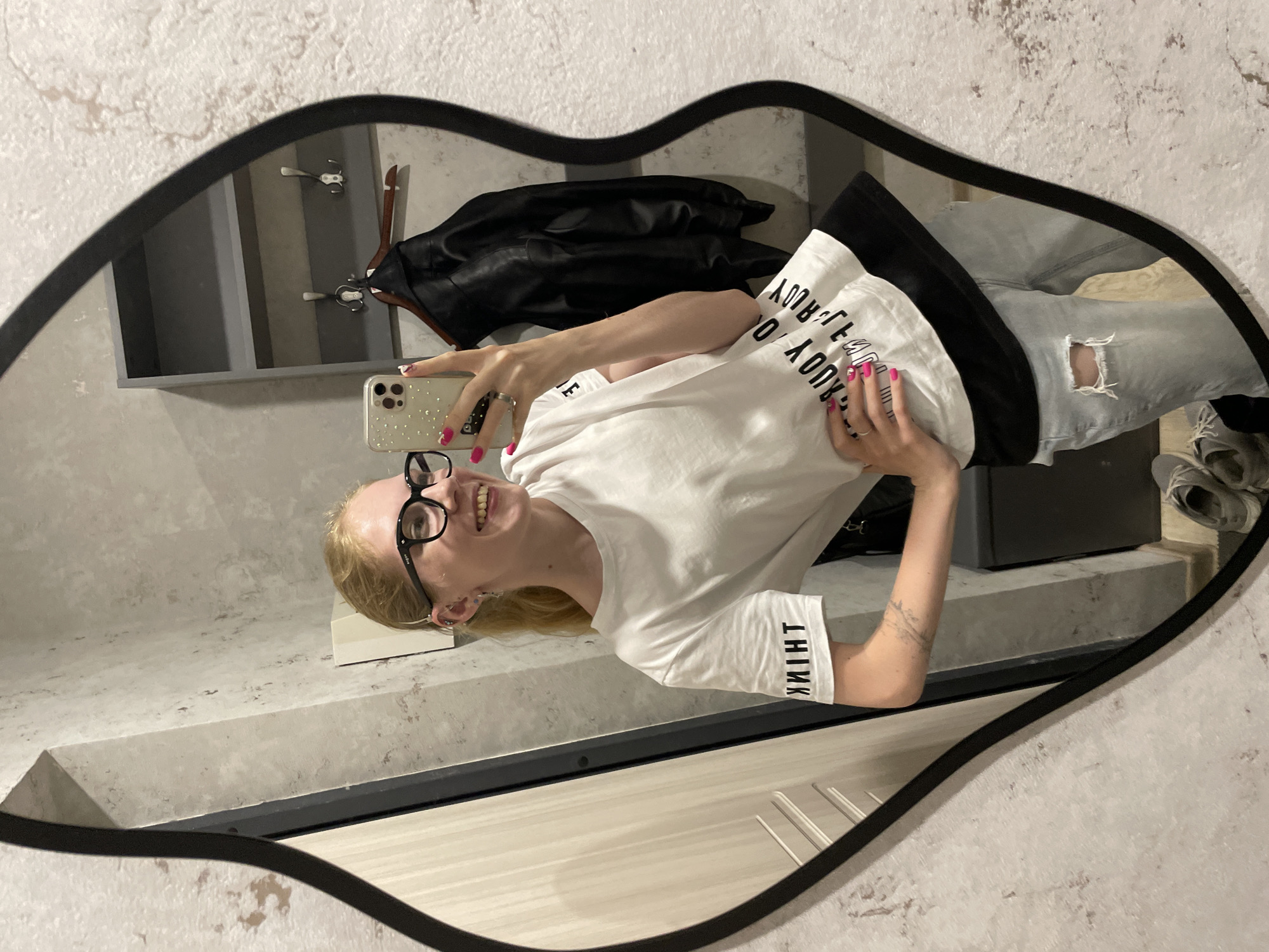 The image shows a person reflected in a decorative mirror, shaped like lips, hanging on a light-colored, textured wall. The person is holding a cell phone, appearing to take a selfie. The person has light blonde hair, wears glasses, and is smiling. The person is wearing a white T-shirt with text and ripped blue jeans. Behind the person, in the reflection, is a closet area with a shelf, a dark jacket hanging on a hanger, and shoes. The location cannot be determined from the image.