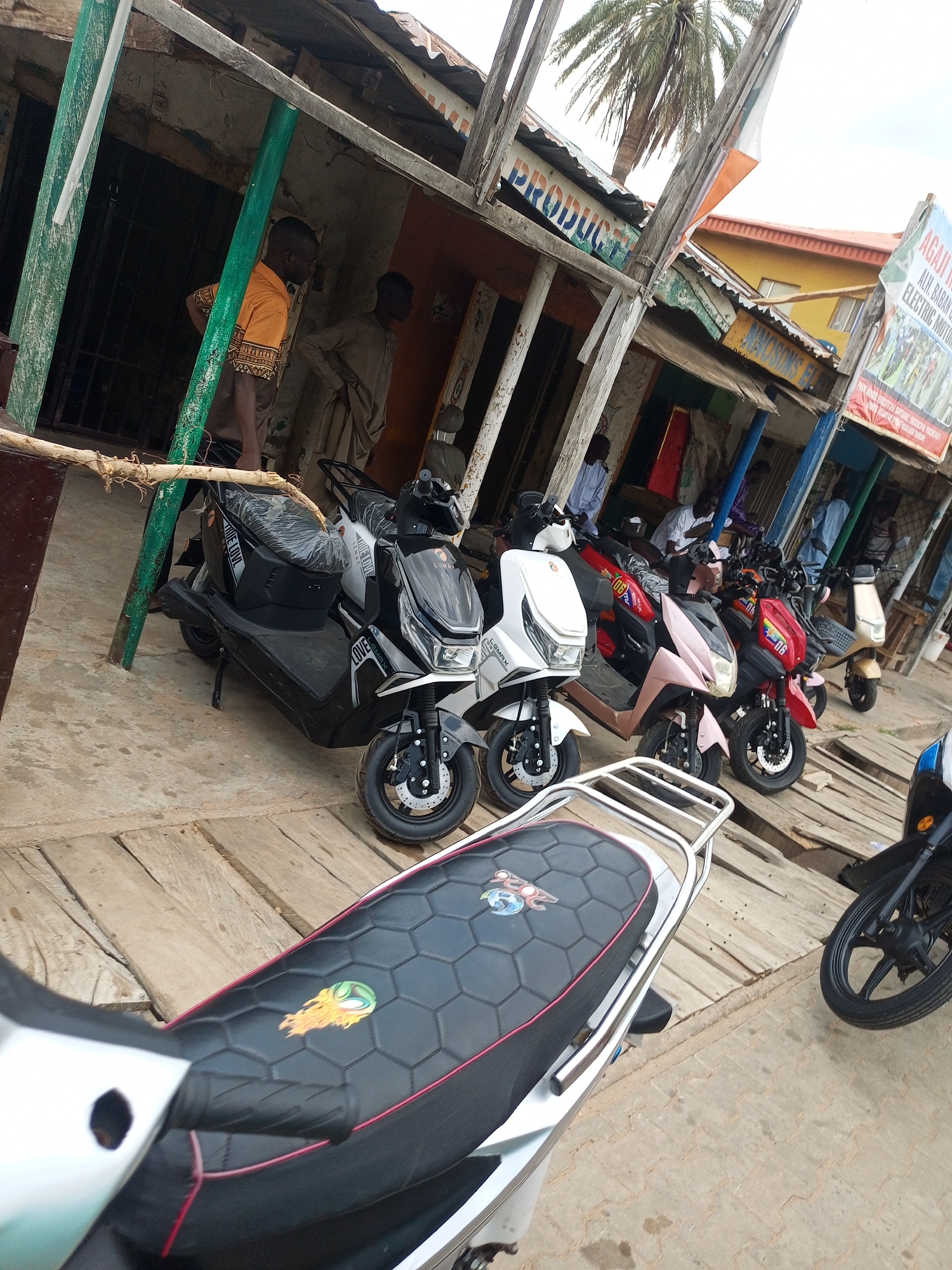 An outdoor street scene in an urban environment features a row of electric scooters parked on a wooden platform in front of a weathered building with a corrugated metal roof. Several people are visible in the background, some standing and others seated within the building's open front. A prominent sign with text, partially obscured by a palm tree, hangs on the building's facade. The scooters are of various colors, including black, white, pink, and red, and appear to be new, with some still having protective plastic on their seats. The ground is a mix of concrete and wood planks. The lighting suggests it is daytime.
